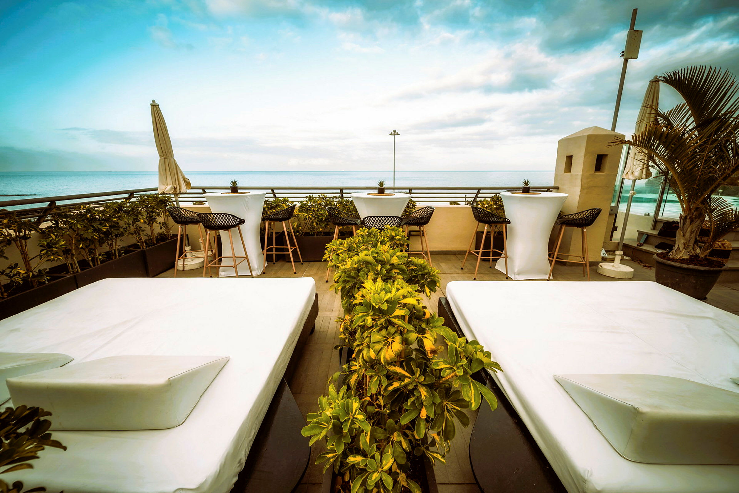 En terrasse med hvite solsenger og små bord med stoler. I bakgrunnen sees havet og blå himmel. (AI-generert tekst)