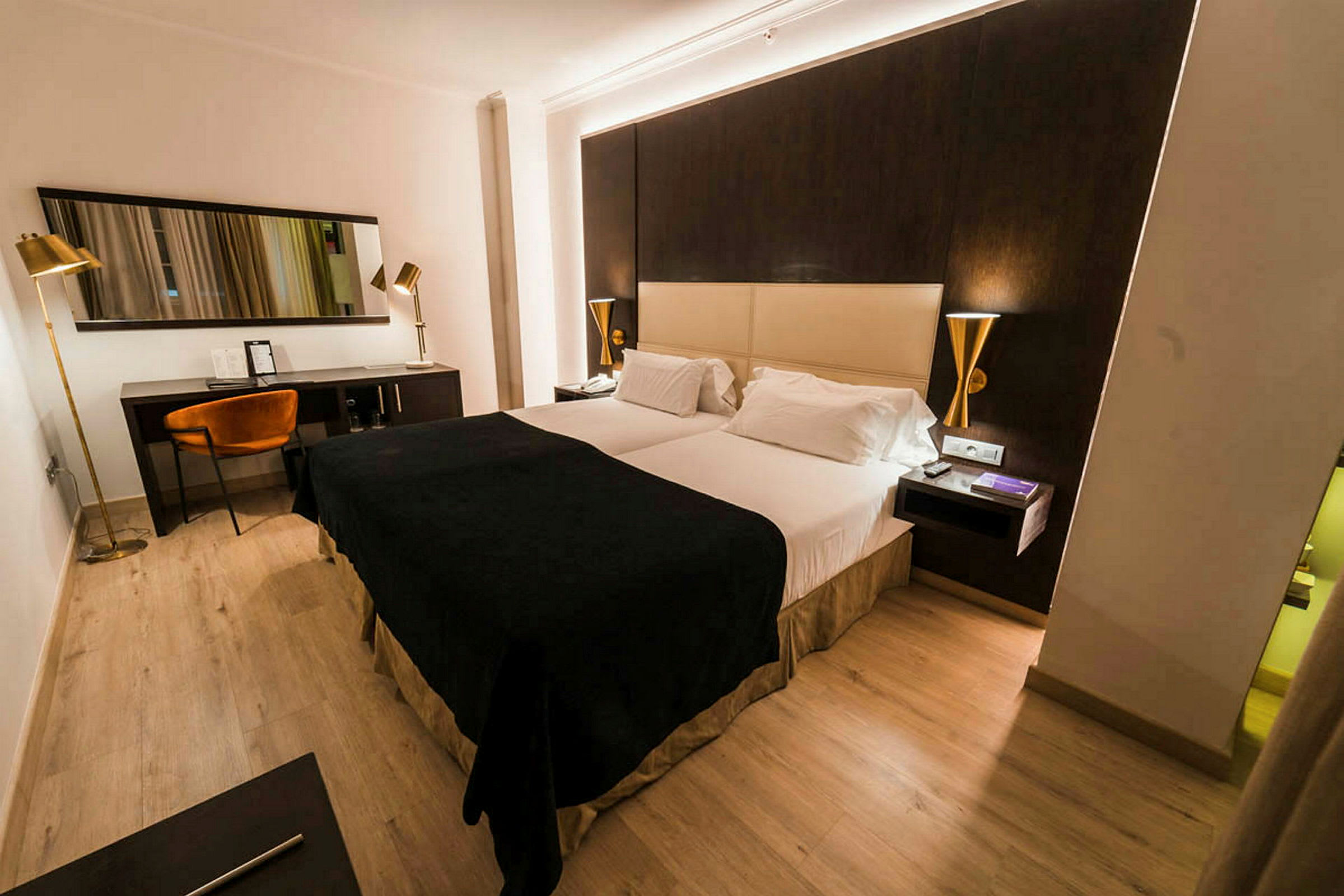 Et hotellrom med en dobbeltseng, mørke og lyse fargetoner på møbler samt en skrivebordskrok med stol og speil. (AI-generert tekst)