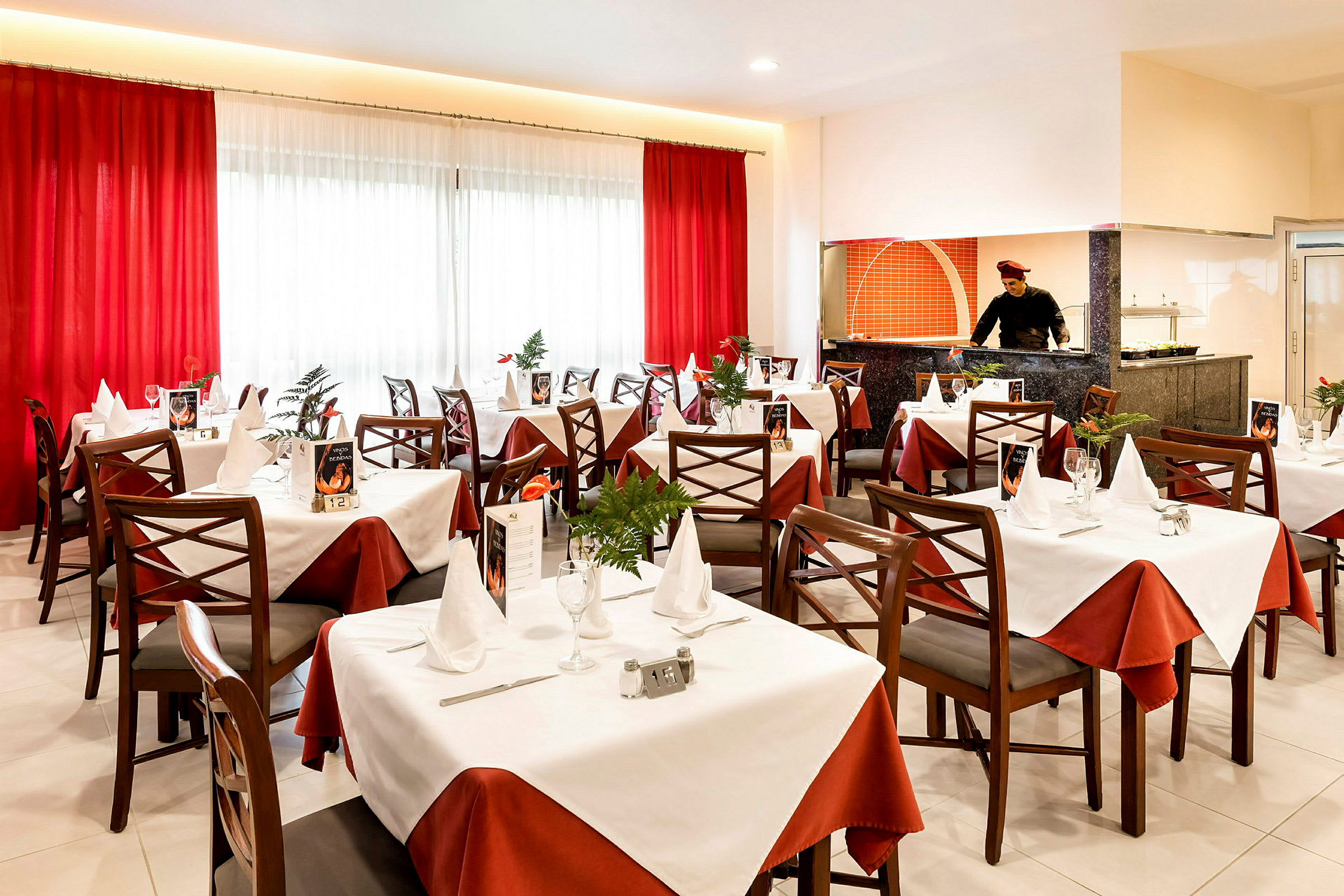 Elegant restaurant med dekkede bord, hvite og røde duker og et åpent kjøkkenområde. (AI-generert tekst)