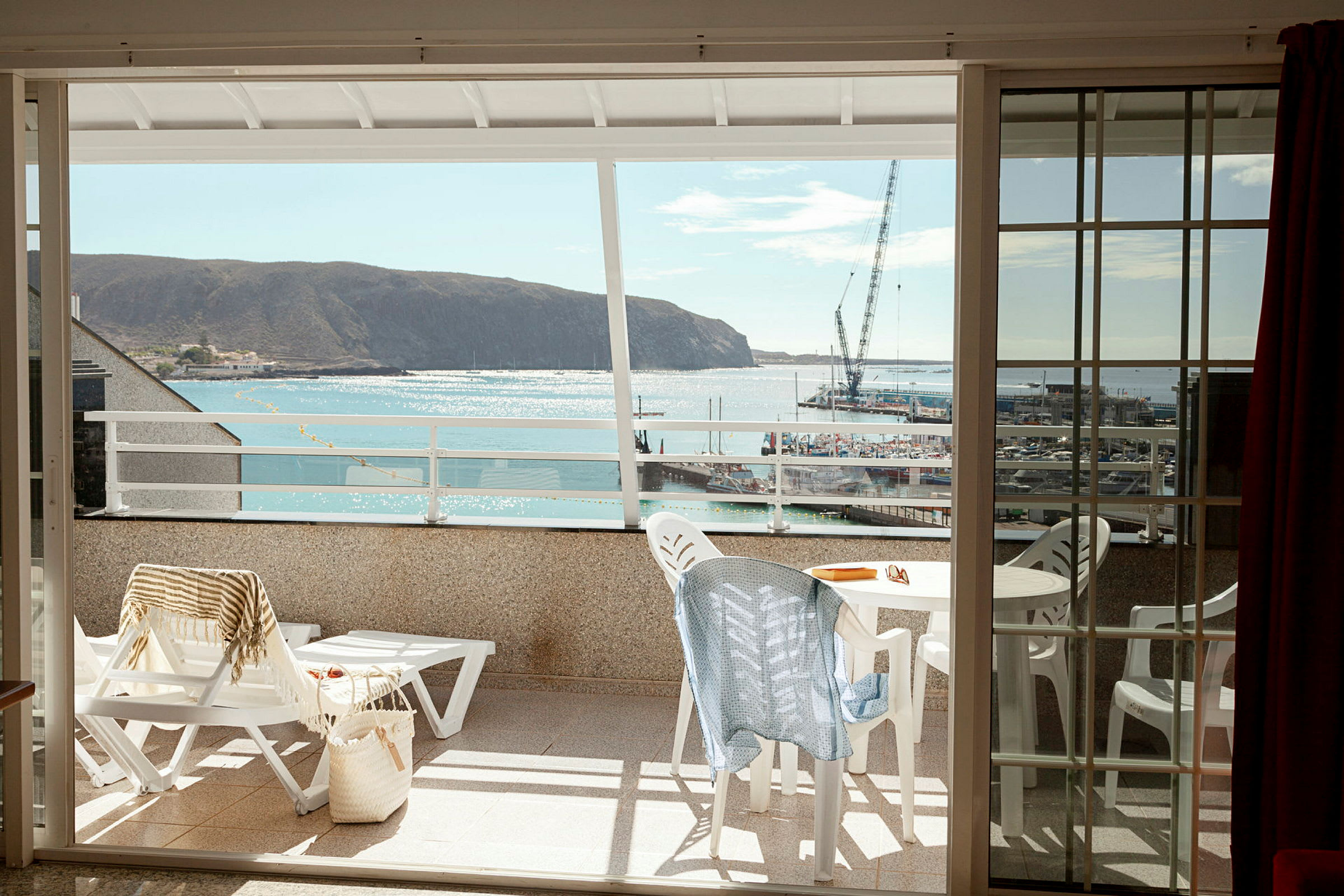 Utsikt fra en balkong mot havet og en liten havn, med utemøbler og solsenger på balkongen. (AI-generert tekst)