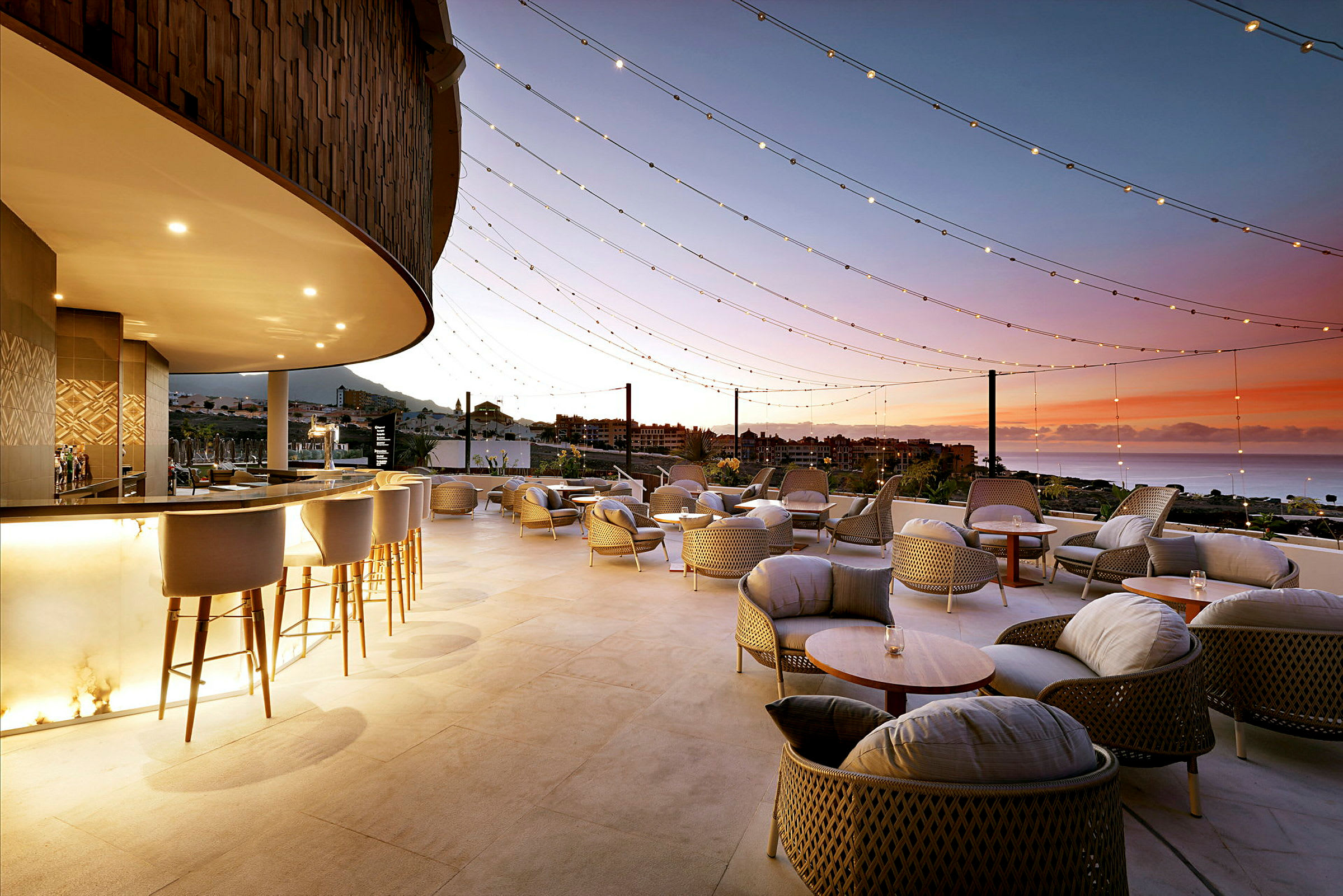 En terrasse med sittegrupper under et tak med lyslenker, med utsikt over havet og en vakker solnedgang. (AI-generert tekst)