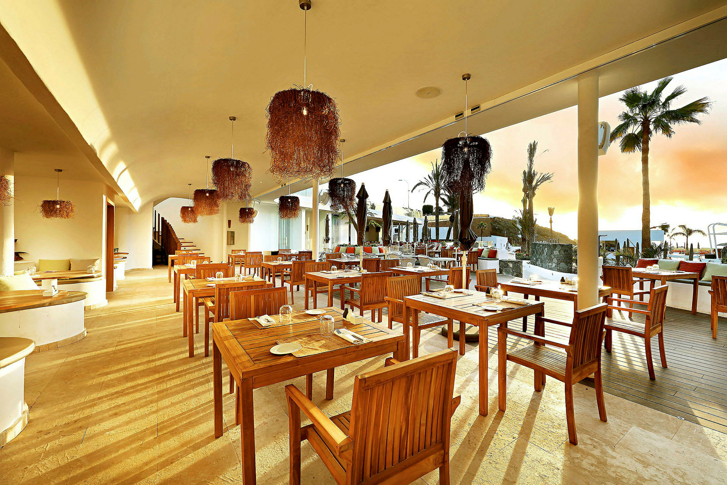 En utendørsrestaurant med trebord og stoler, moderne innredning med hengende lamper, solnedgang i bakgrunnen. (AI-generert tekst)