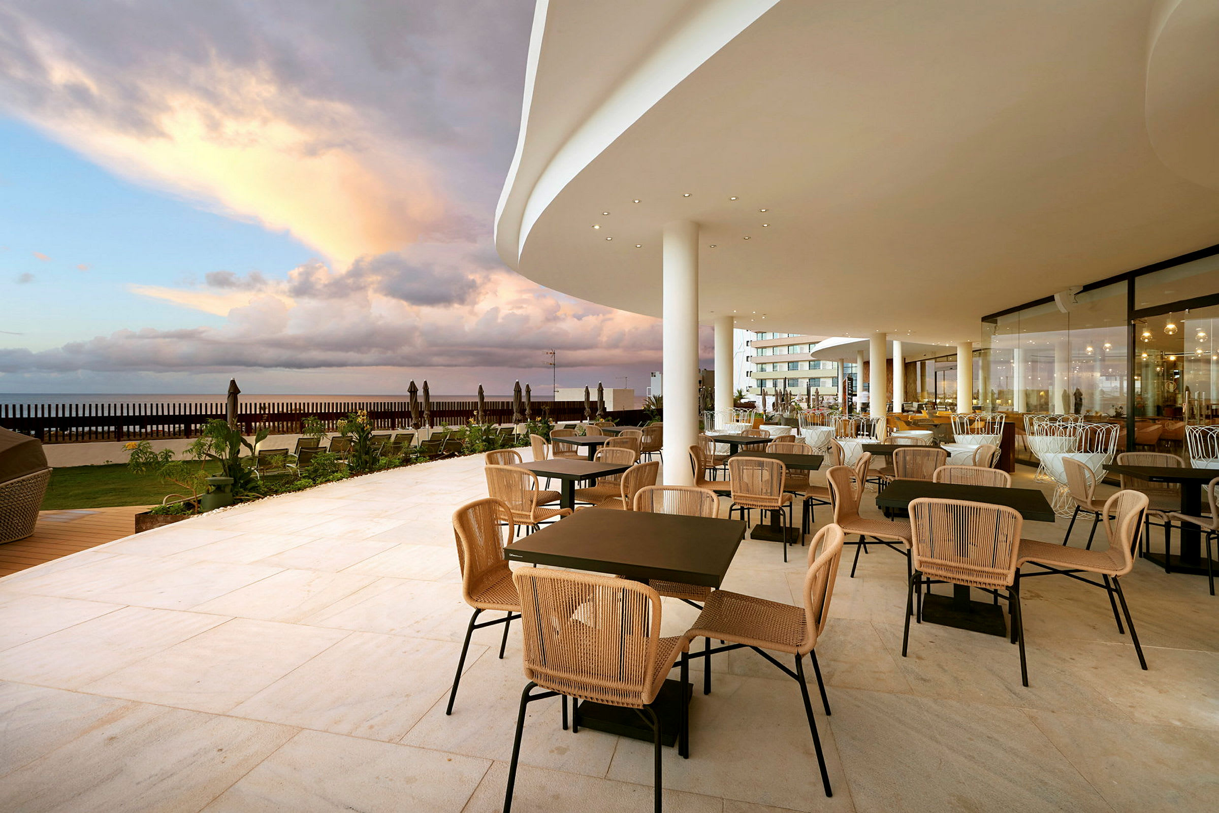 En restaurantterrasse med bord og stoler samt utsikt over havet under en skyet himmel. (AI-generert tekst)