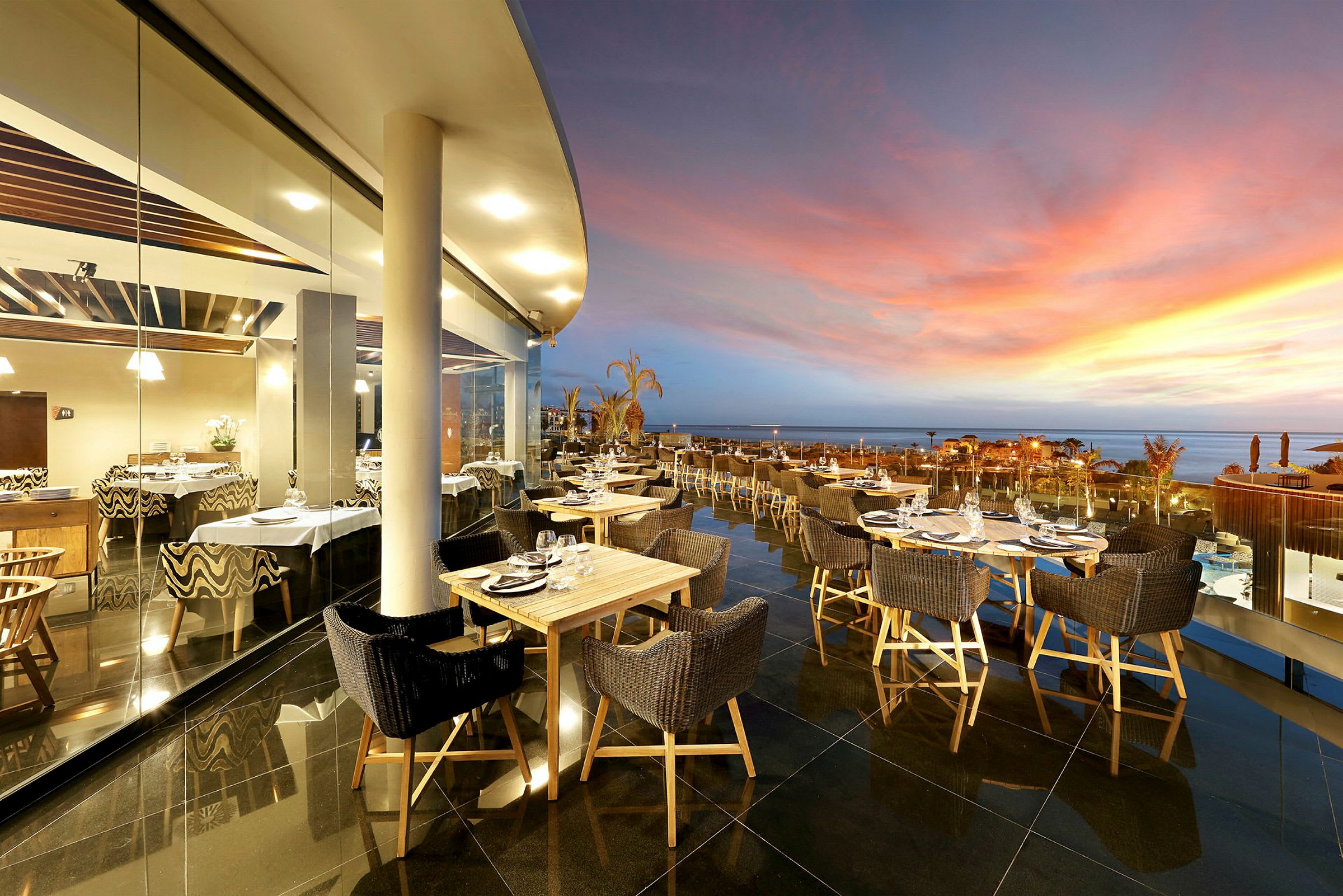 En utendørs restaurant med bord og stoler ved en glassvegg med utsikt over en havsbelyst solnedgang. (AI-generert tekst)