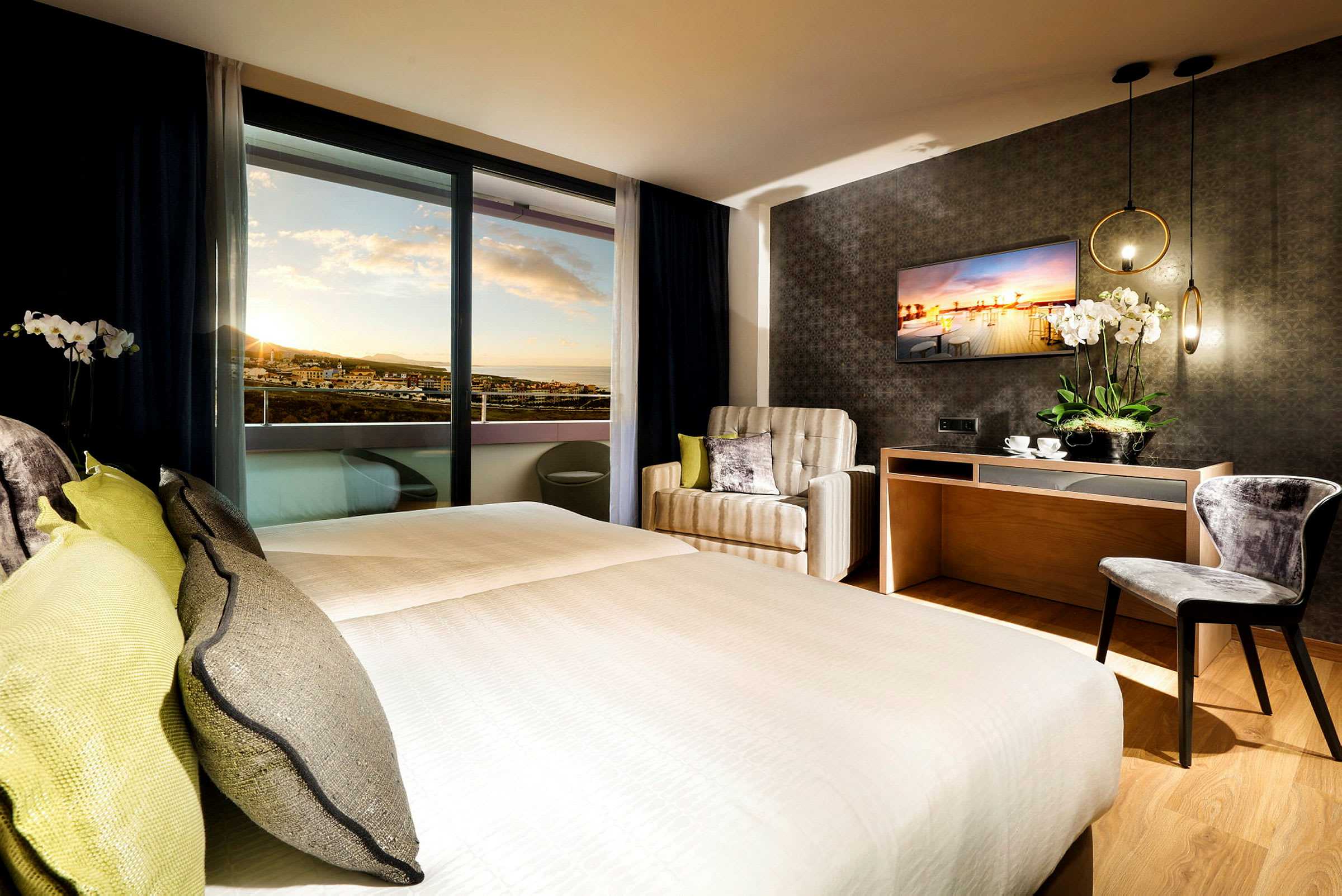 Hotellrom med dobbeltseng, lenestol, skrivebord og utsikt mot solnedgangen. (AI-generert tekst)