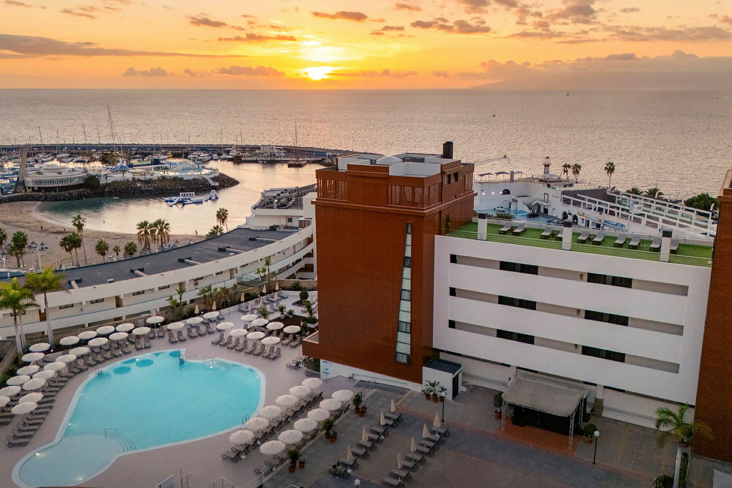 Bilde av et hotell med bassengområde i forgrunnen og en solnedgang over en marina i bakgrunnen. (AI-generert tekst)