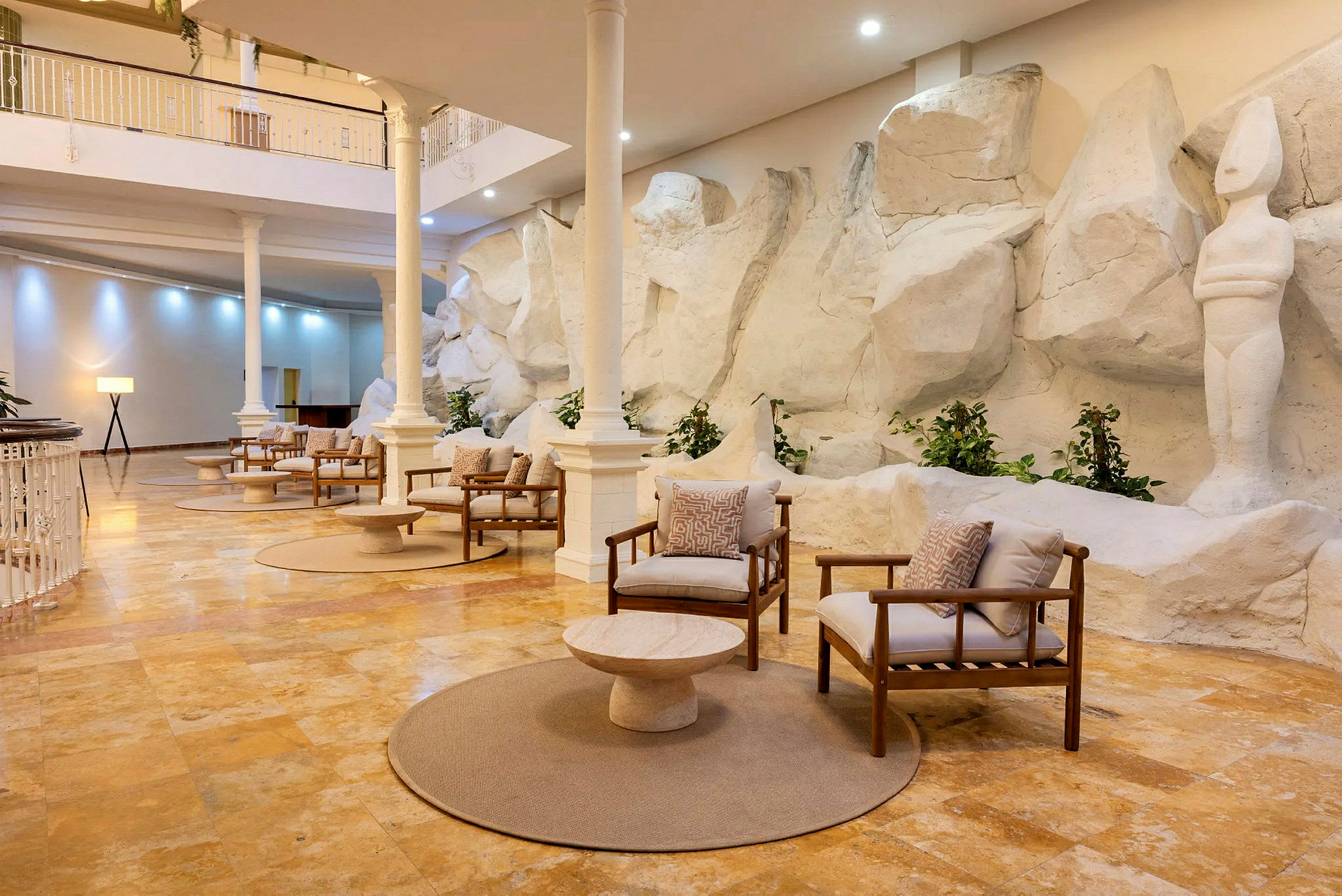 En elegant lounge med trestoler og puter, plassert på et steingulv, omgitt av hvite skulpturer og steinvegger, under et åpent innendørsmiljø. (AI-generert tekst)
