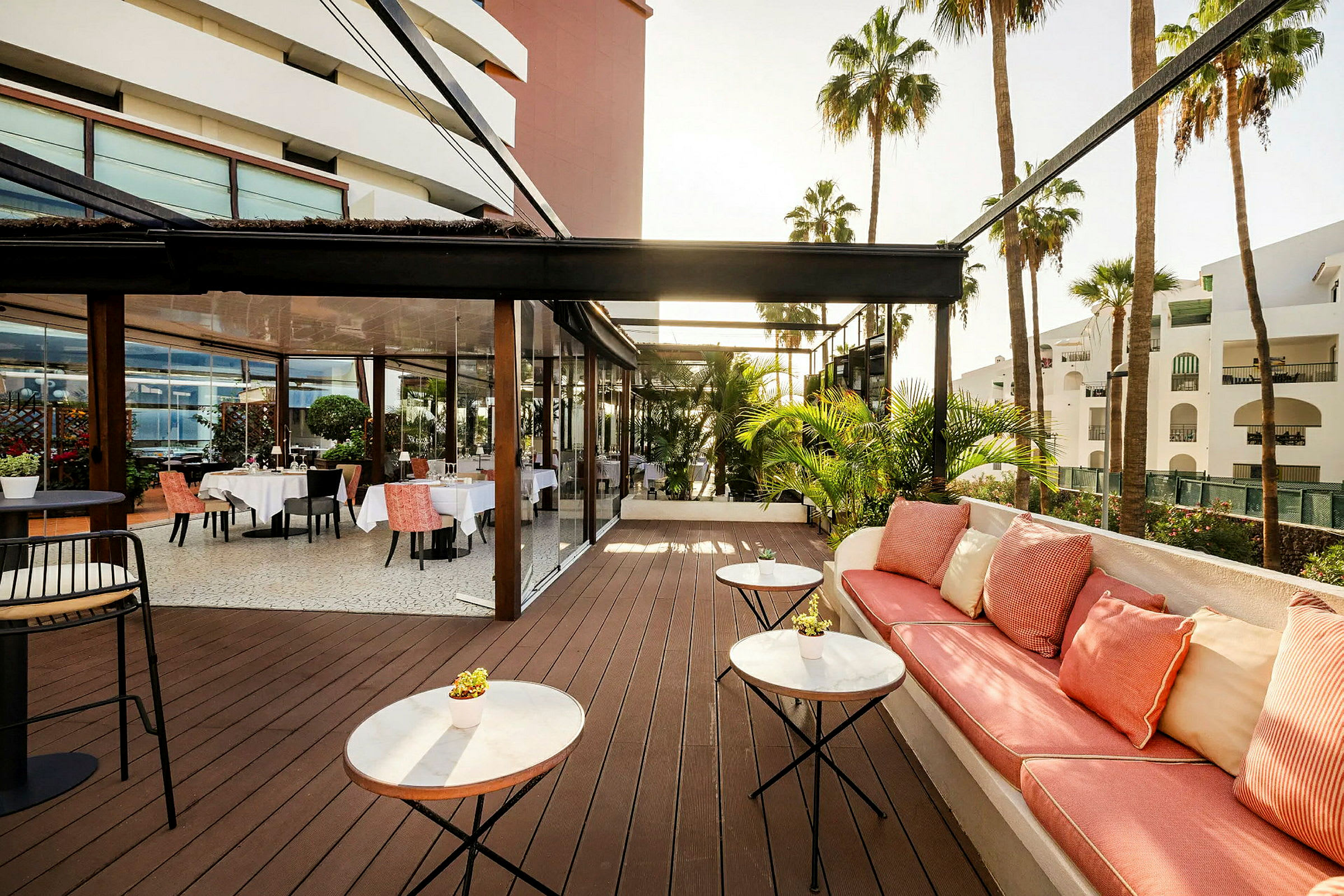 En utendørs terrasse med bord og stoler samt en sitteplass med puter. Palmer og hvite bygninger er synlige i bakgrunnen. (AI-generert tekst)