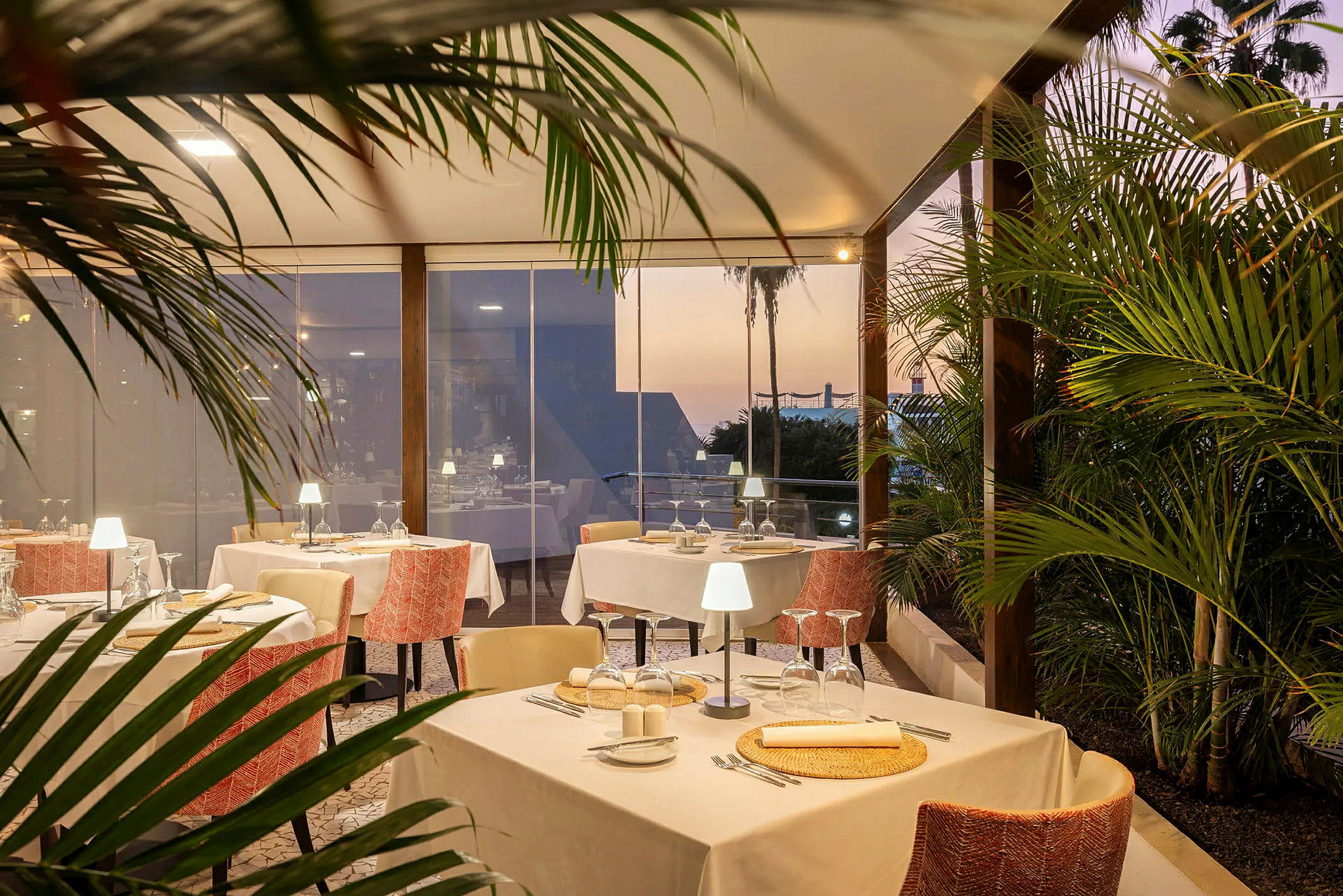 En elegant restaurant med dekkede bord inne og omgitt av grøntområder, med solnedgang i bakgrunnen. (AI-generert tekst)