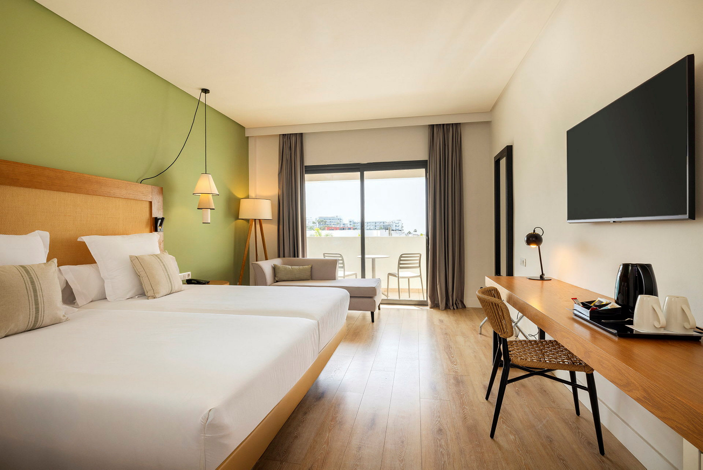 Ett hotellrom med to senger, grønne vegger, skrivebord og sittegruppe ved et vindu med utsikt til balkongen. (AI-generert tekst)