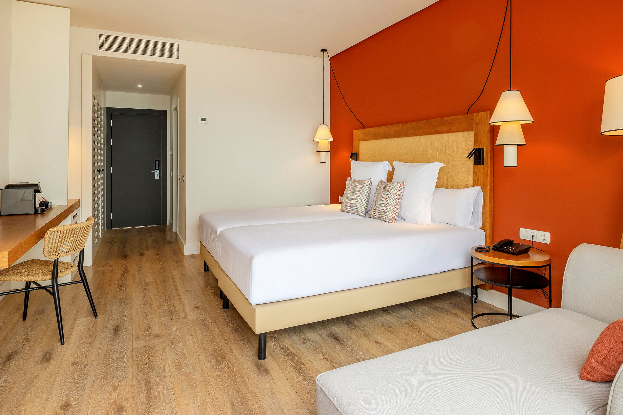 Et hotellrom med en oransje vegg, en dobbeltseng, skrivebord og stoler. (AI-generert tekst)