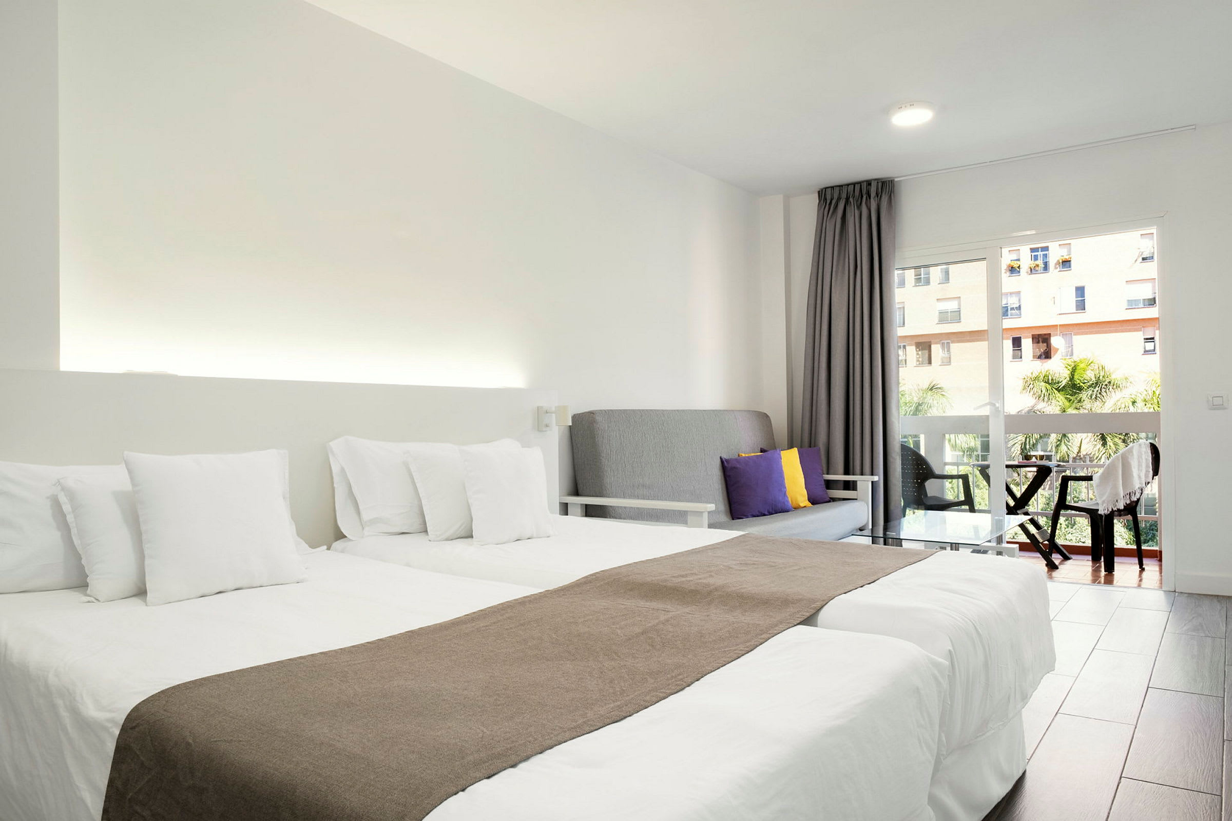 Et hotellrom med to enkeltsenger med hvitt sengetøy, en grå sofa med fargede puter og en balkong med utsikt. (AI-generert tekst)