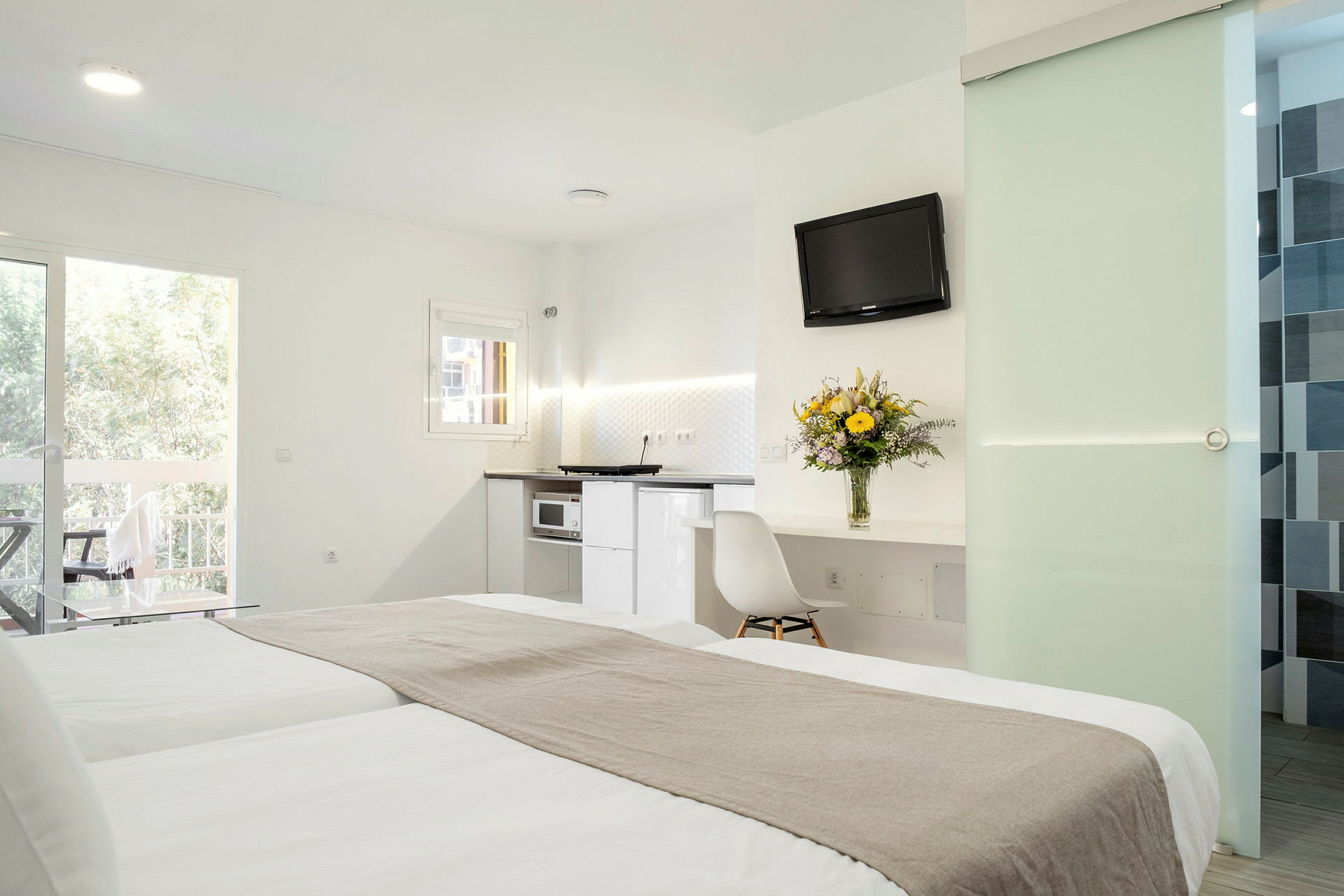 Et hotellrom med en dobbeltseng, lys innredning, et kjøkken, en balkong og et bord med en blomsterbukett. (AI-generert tekst)