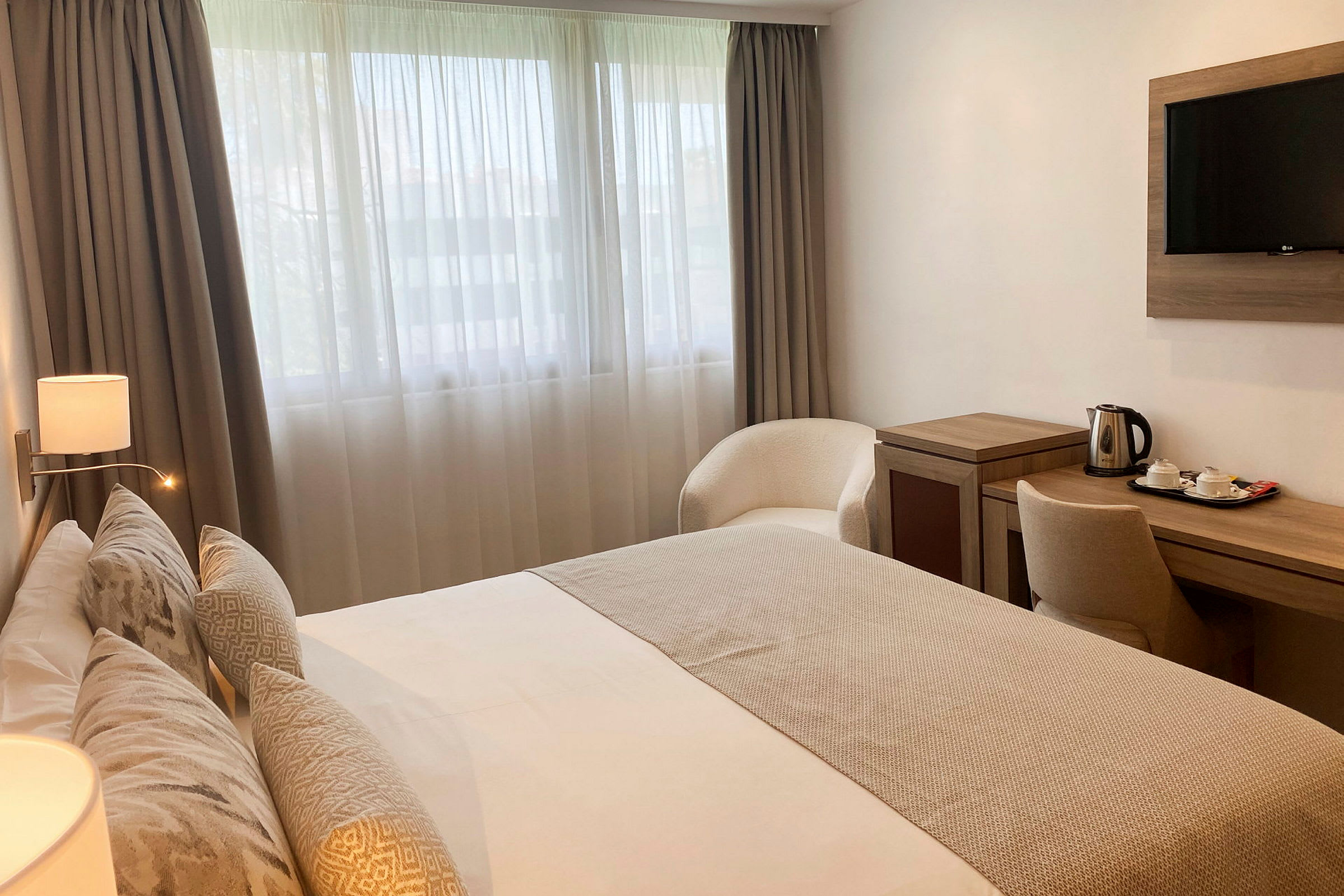 Hotellrom med en dobbeltseng, skrivebord med vannkoker og tefatsett, samt en lenestol plassert nær vinduet. (AI-generert tekst)