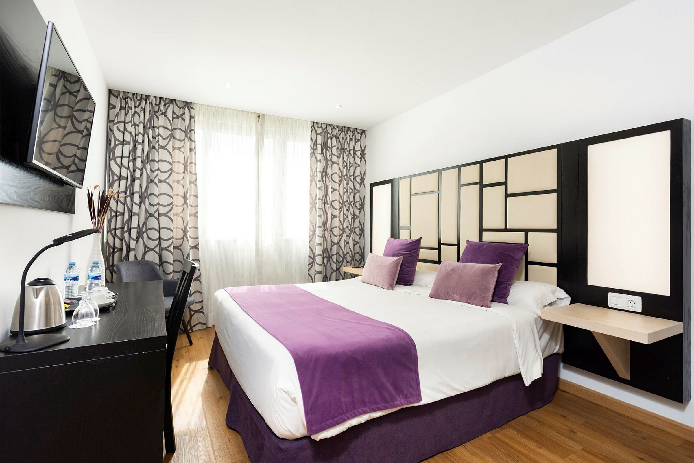 Et hotellrom med en dobbeltseng, dekorert med lilla puter og sengetepper og en moderne veggdesign. (AI-generert tekst)