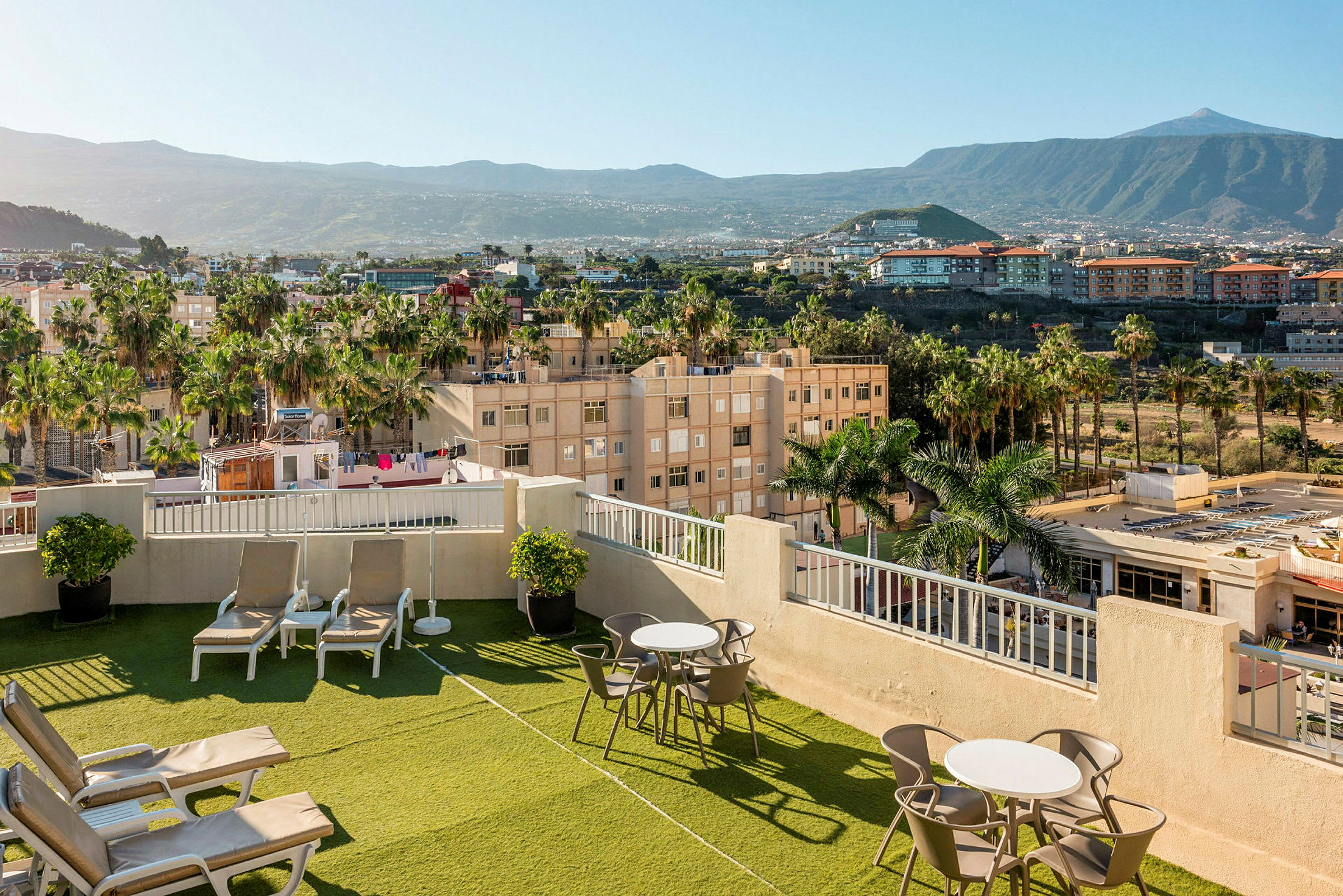 Bilde av en hotelltakterrasse med solsenger og bord, omgitt av grønne områder og palmer, med utsikt over omliggende byområde og fjell. (AI-generert tekst)