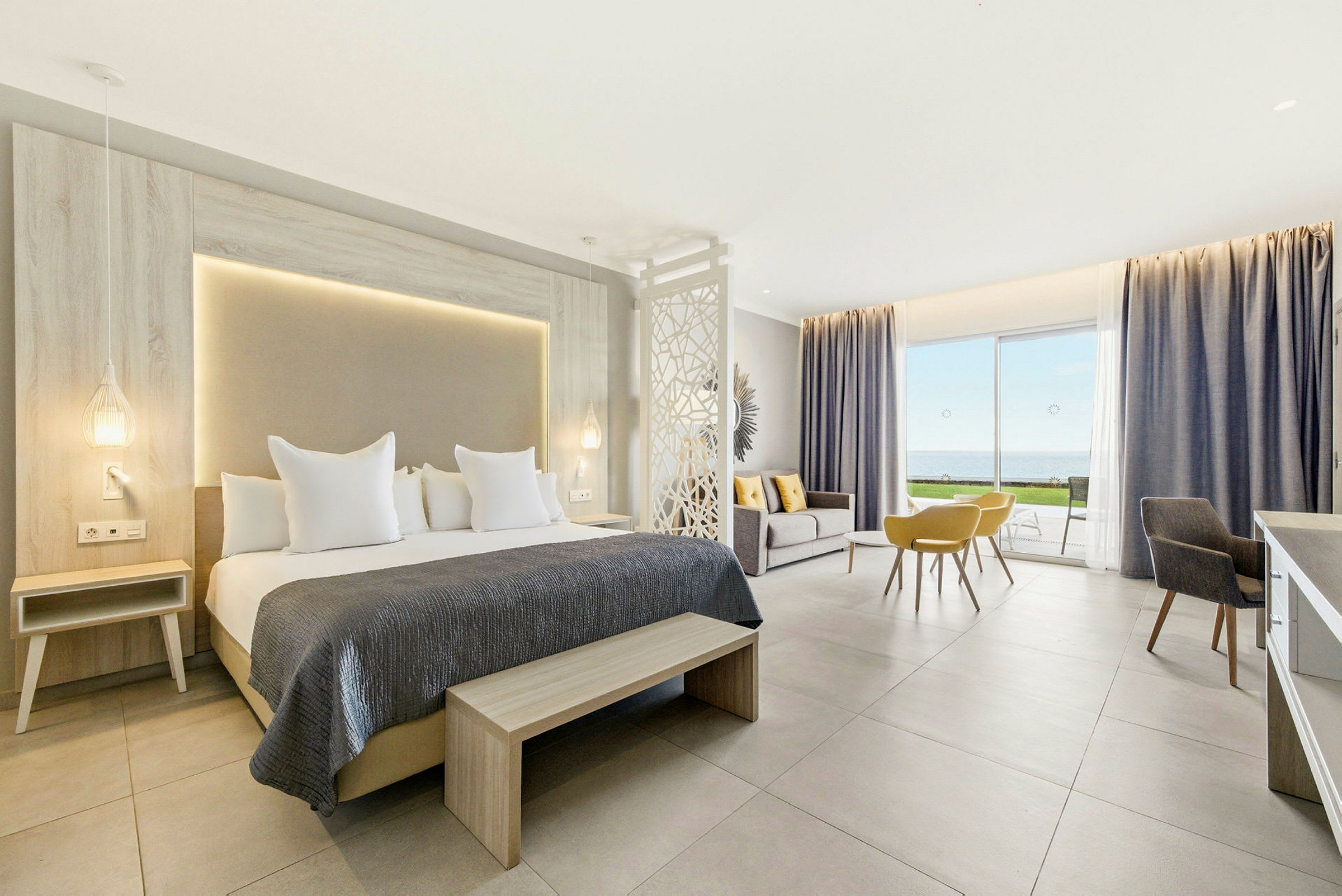 Et moderne hotellrom med dobbeltseng, sitteområde og utsikt over havet gjennom store glassdører. (AI-generert tekst)