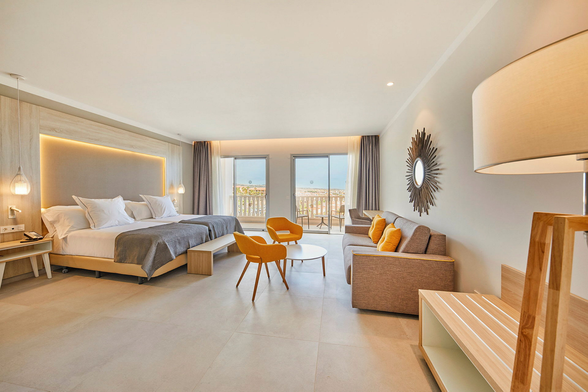 Et moderne hotellrom med dobbeltseng, sittegruppe med oransje stoler, en sofa og en balkong med utsikt mot havet. (AI-generert tekst)