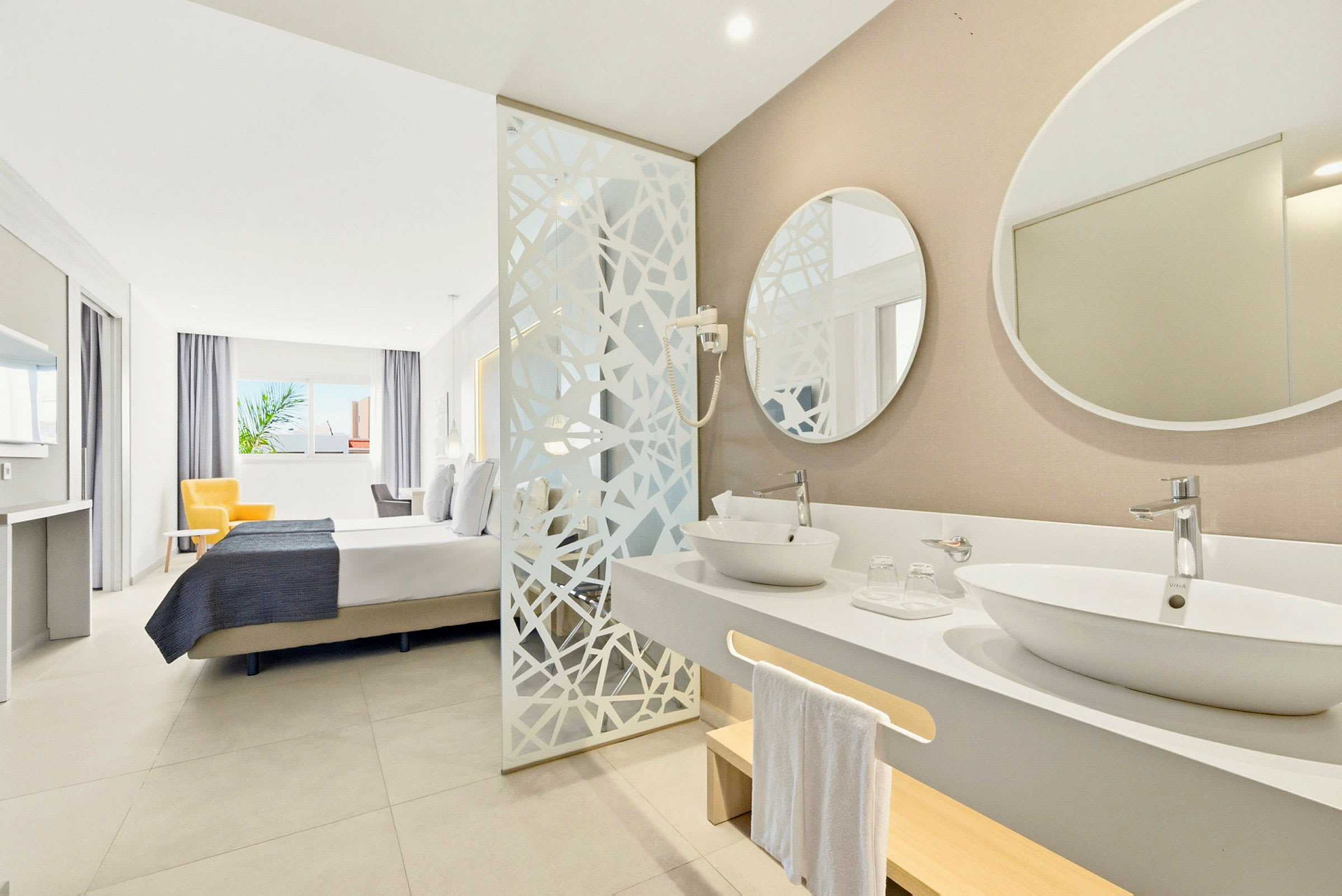 Et hotellrom med bad og sovested. Badet har to vasker og et speil. Sovestedet har en seng og en gul lenestol ved et stort vindu. (AI-generert tekst)