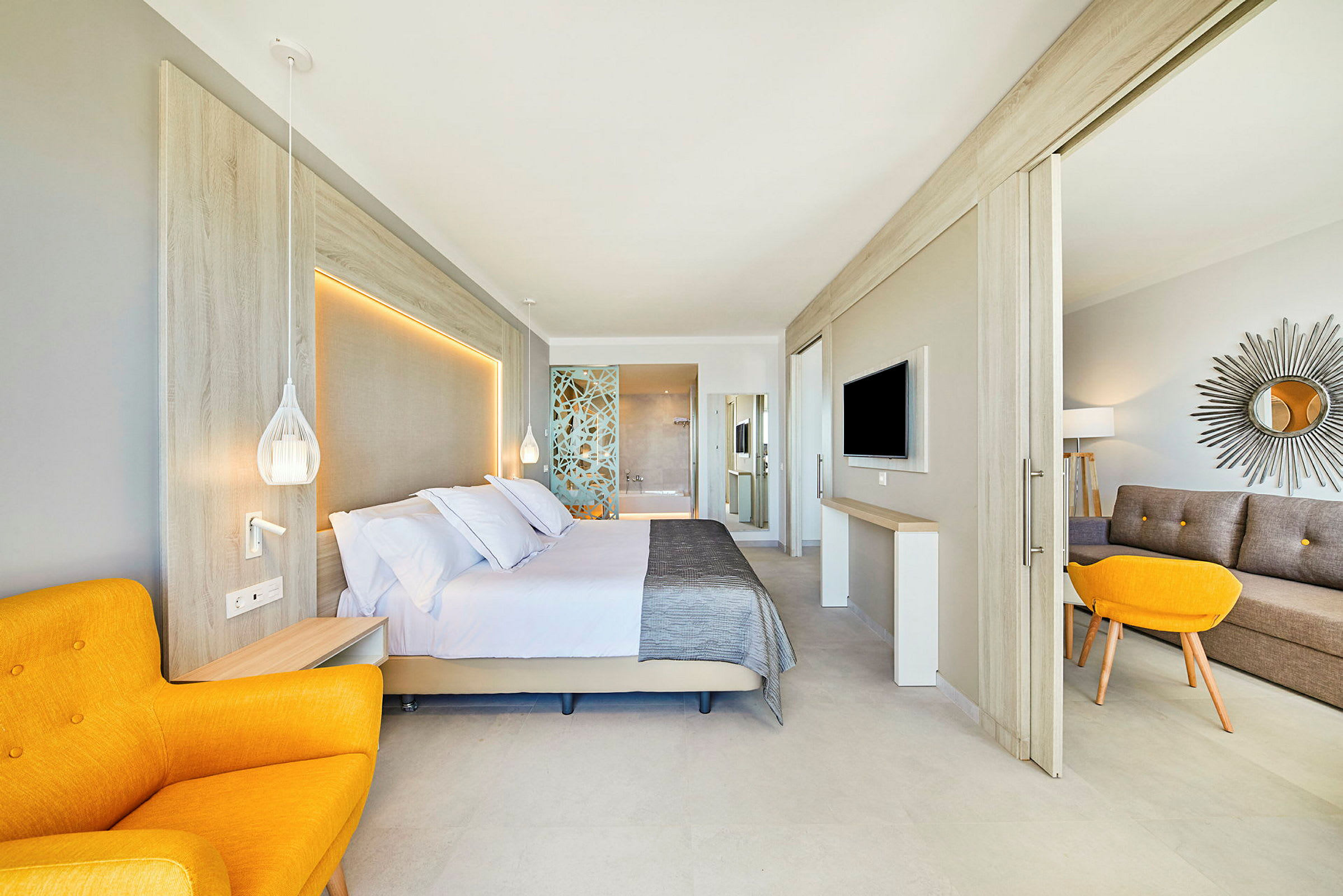 Et moderne hotellrom med en stor seng, en gul lenestol, samt en sittegruppe og flatskjerm-TV. Innredningen er lys og stilren. (AI-generert tekst)