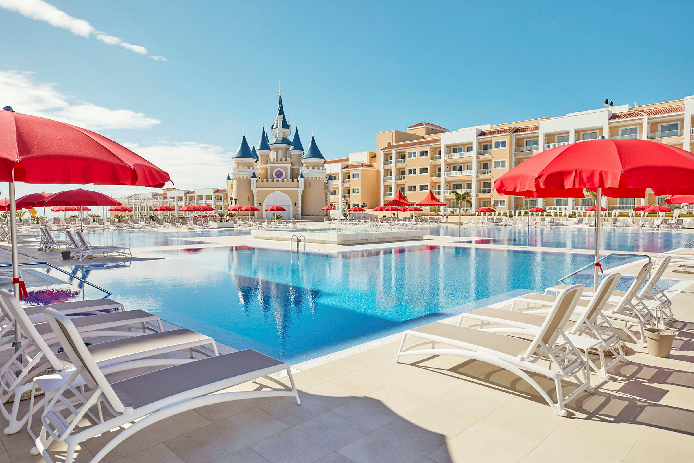 Et stort basseng med solsenger og røde parasoller, samt en bygning med et slottslignende utseende i bakgrunnen. (AI-generert tekst)