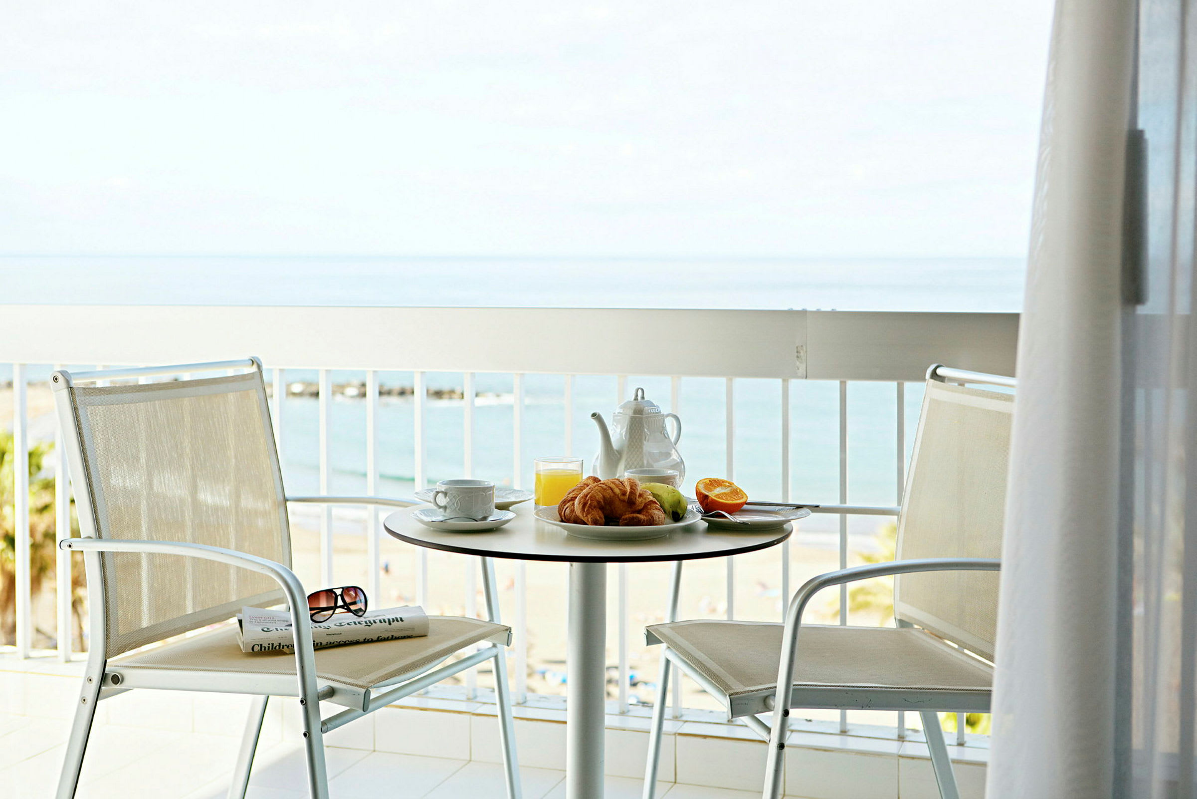 En balkong med utsikt over havet og en strand. På et bord står et brett med frokost som består av croissanter, en halv appelsin, eple, appelsinjuice og en tekanne. (AI-generert tekst)