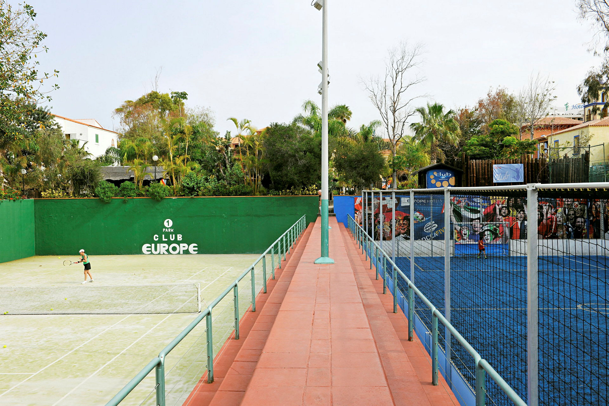 En grønn tennisbane til venstre og en blå padelbane til høyre, omgitt av trær og bygninger. (AI-generert tekst)