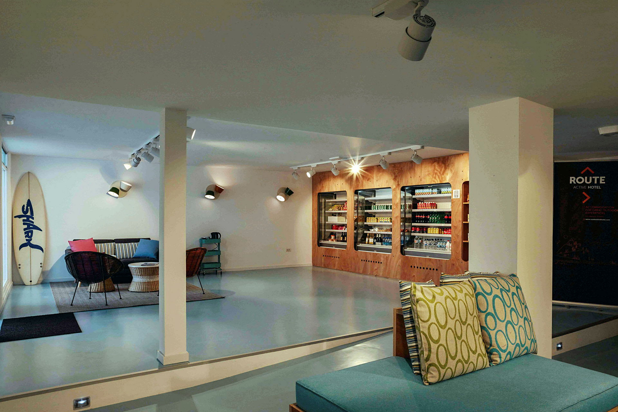 Interiør av en moderne lounge med sofaer, bord og kjøleskap fylt med varer, samt et surfebrett som står lent mot veggen. (AI-generert tekst)