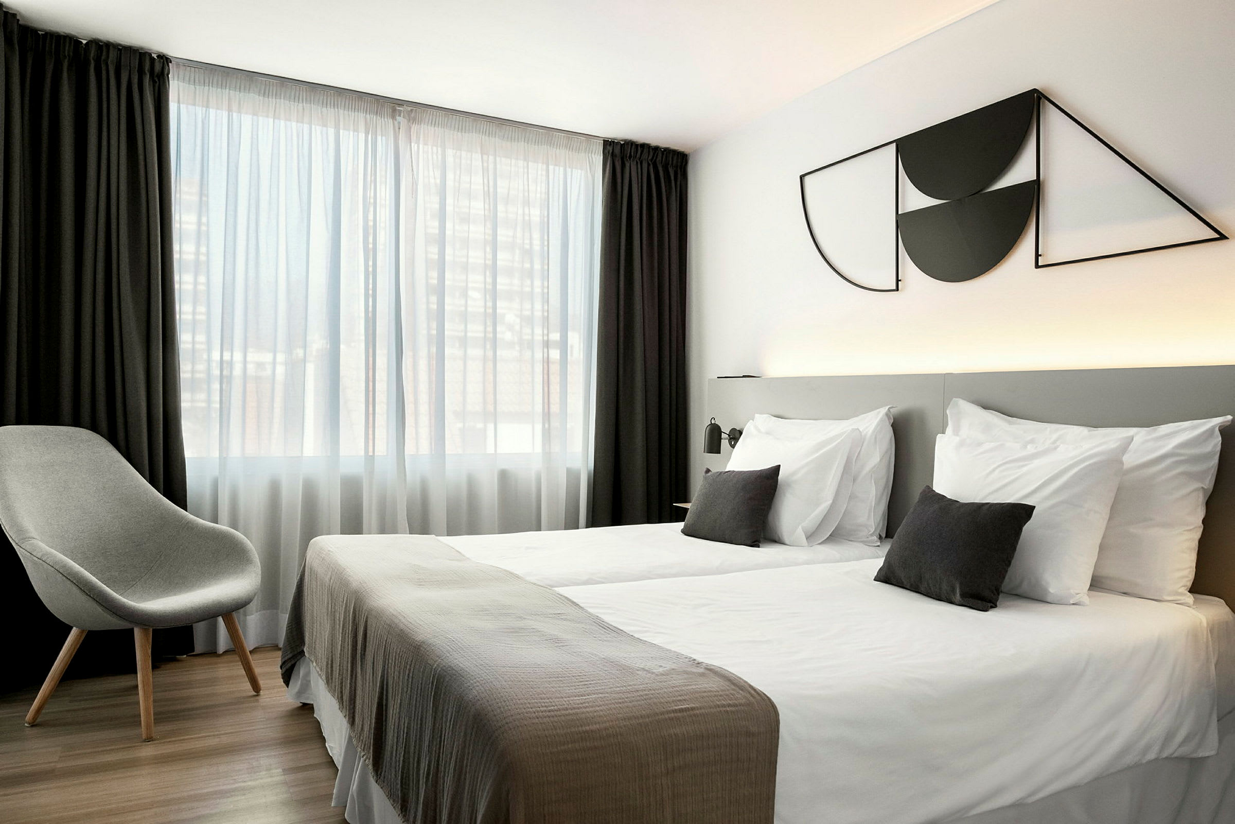 Et hotellrom med to enkeltsenger, en grå stol og et stort vindu med hvite og mørke gardiner. (AI-generert tekst)