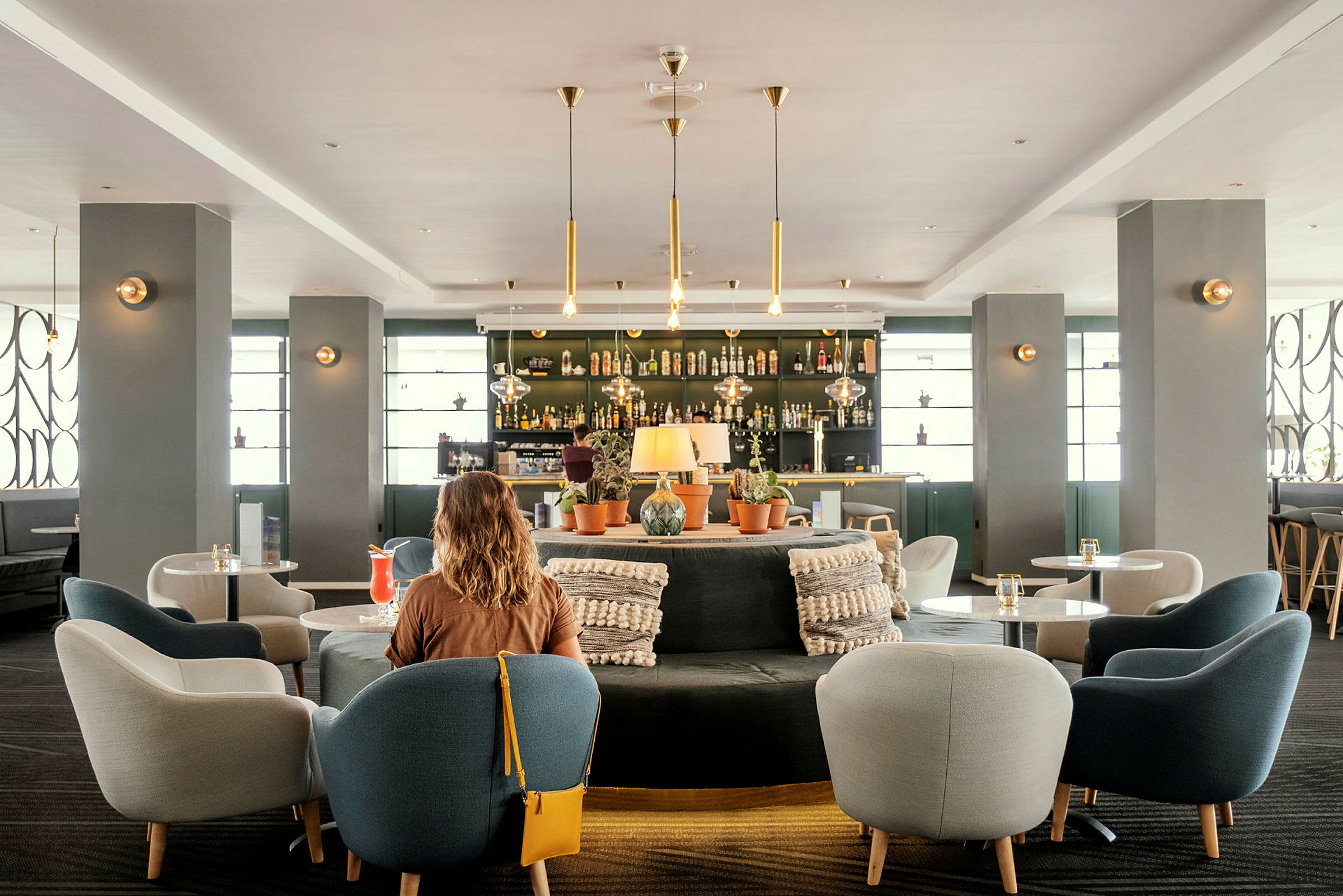 Interiør av et moderne hotellbarområde med sitteplasser og dekorasjoner. En kvinne med ryggen til kameraet i forgrunnen. (AI-generert tekst)