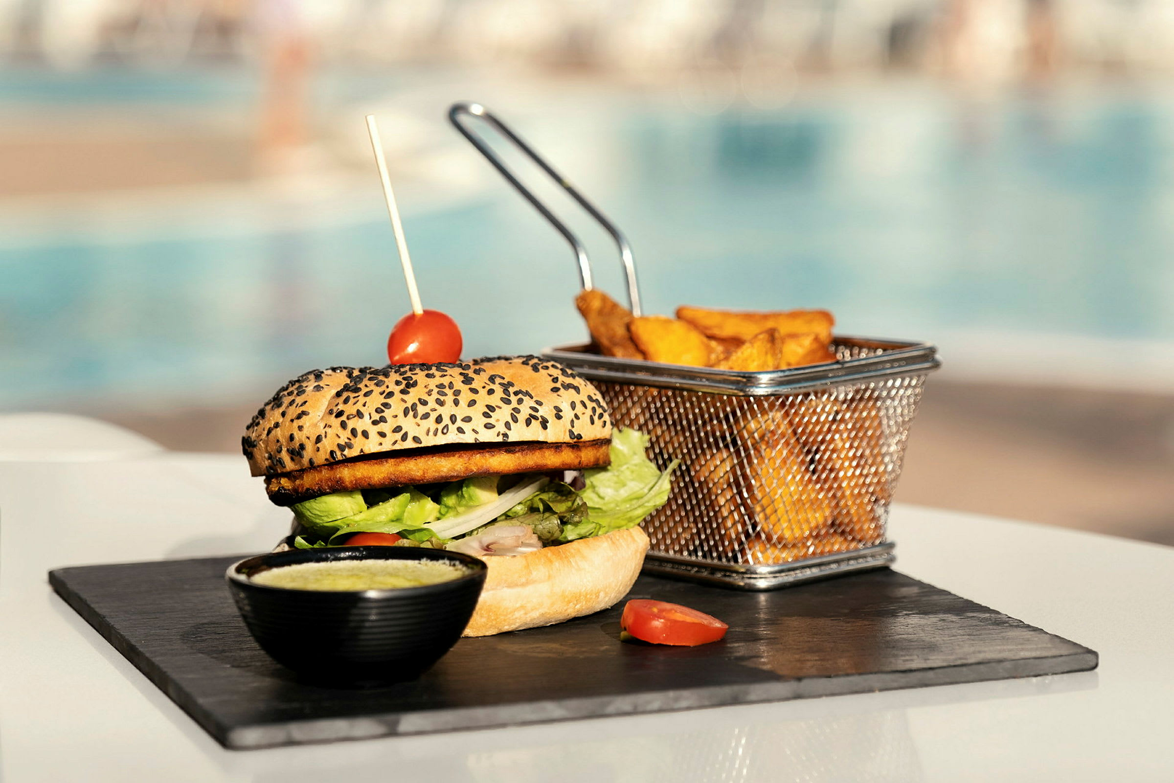 En hamburger med tomatdekorasjon, ved siden av pommes frites i en metallkurv, servert på en steinplate. (AI-generert tekst)
