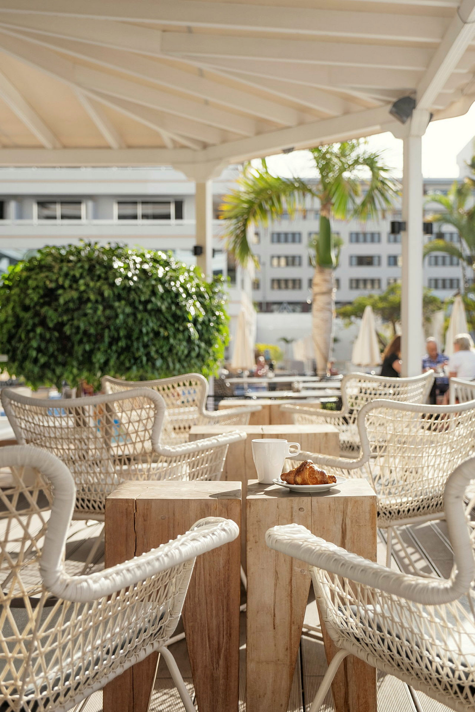 Et kafébord med to trestoler under en pergola, med en kaffekopp og en croissant på bordet. Bakgrunnen inkluderer tropiske planter og andre bord. (AI-generert tekst)