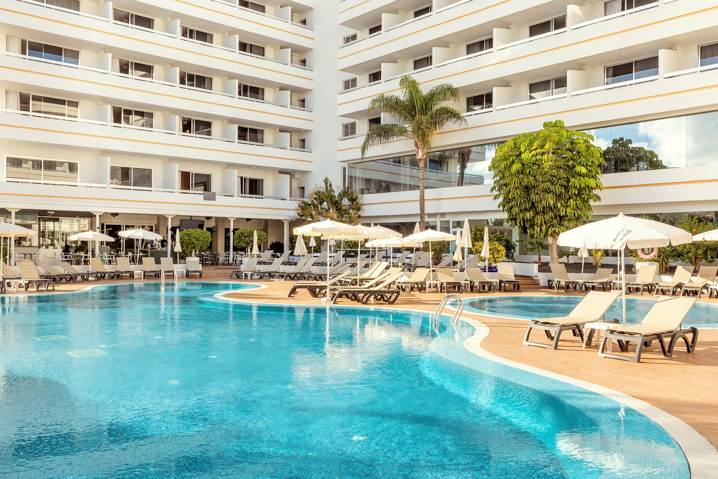 Bilde av et hotellbasseng med solsenger og parasoller ved siden av og et fleretasjes hotell i bakgrunnen. (AI-generert tekst)