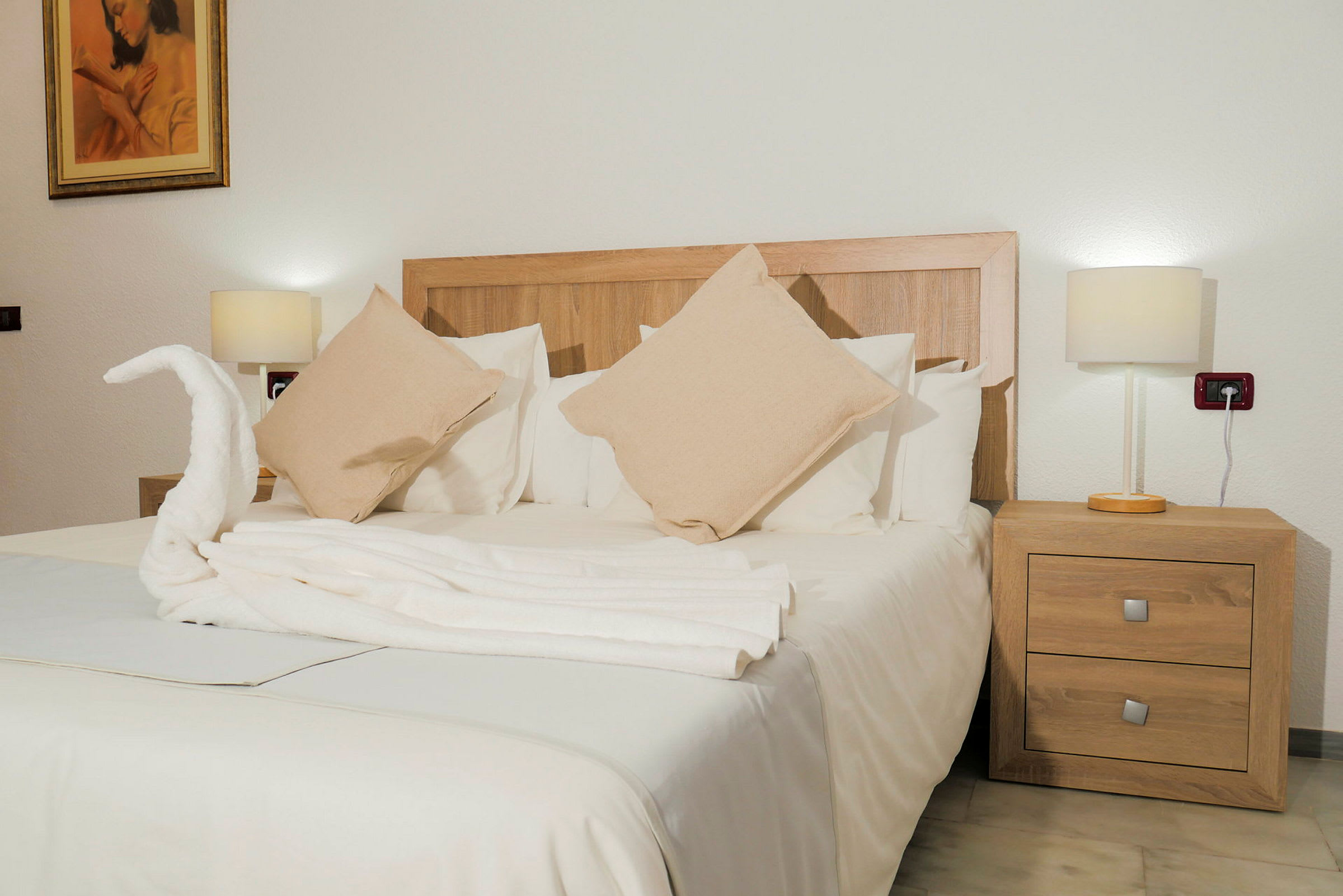 Et hotellrom med en dobbeltseng oppredd med hvitt sengetøy og beige puter, omgitt av to nattbord med lamper. (AI-generert tekst)