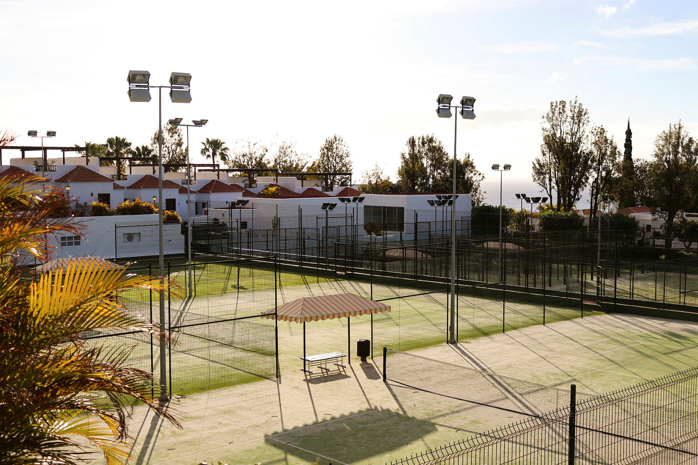 Bilde av en padeltennisbane omgitt av gjerder, med utsikt over en bygning med hvite vegger og røde tak. (AI-generert tekst)