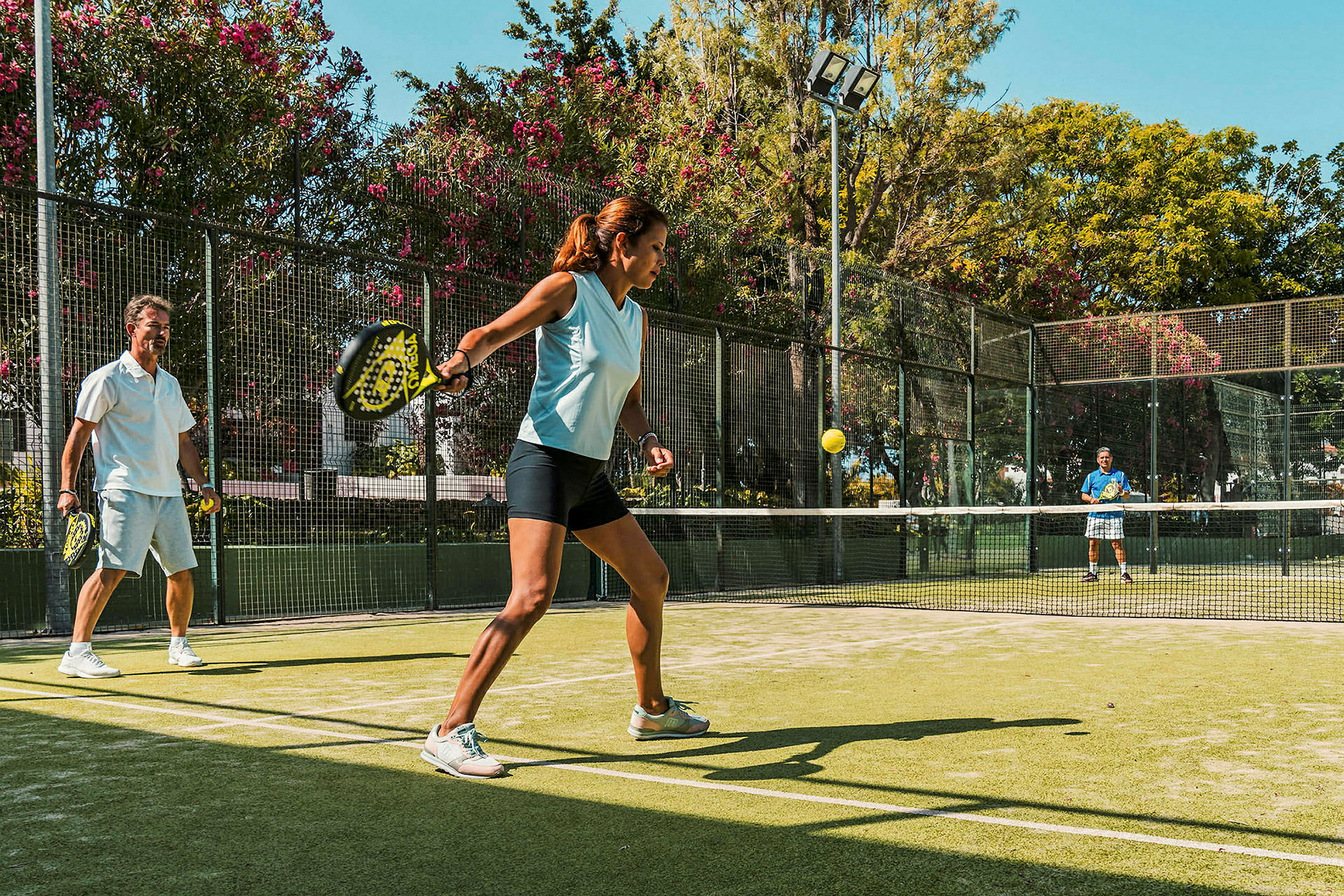 Personer spiller padel på en utendørs bane omgitt av grønn vegetasjon. (AI-generert tekst)