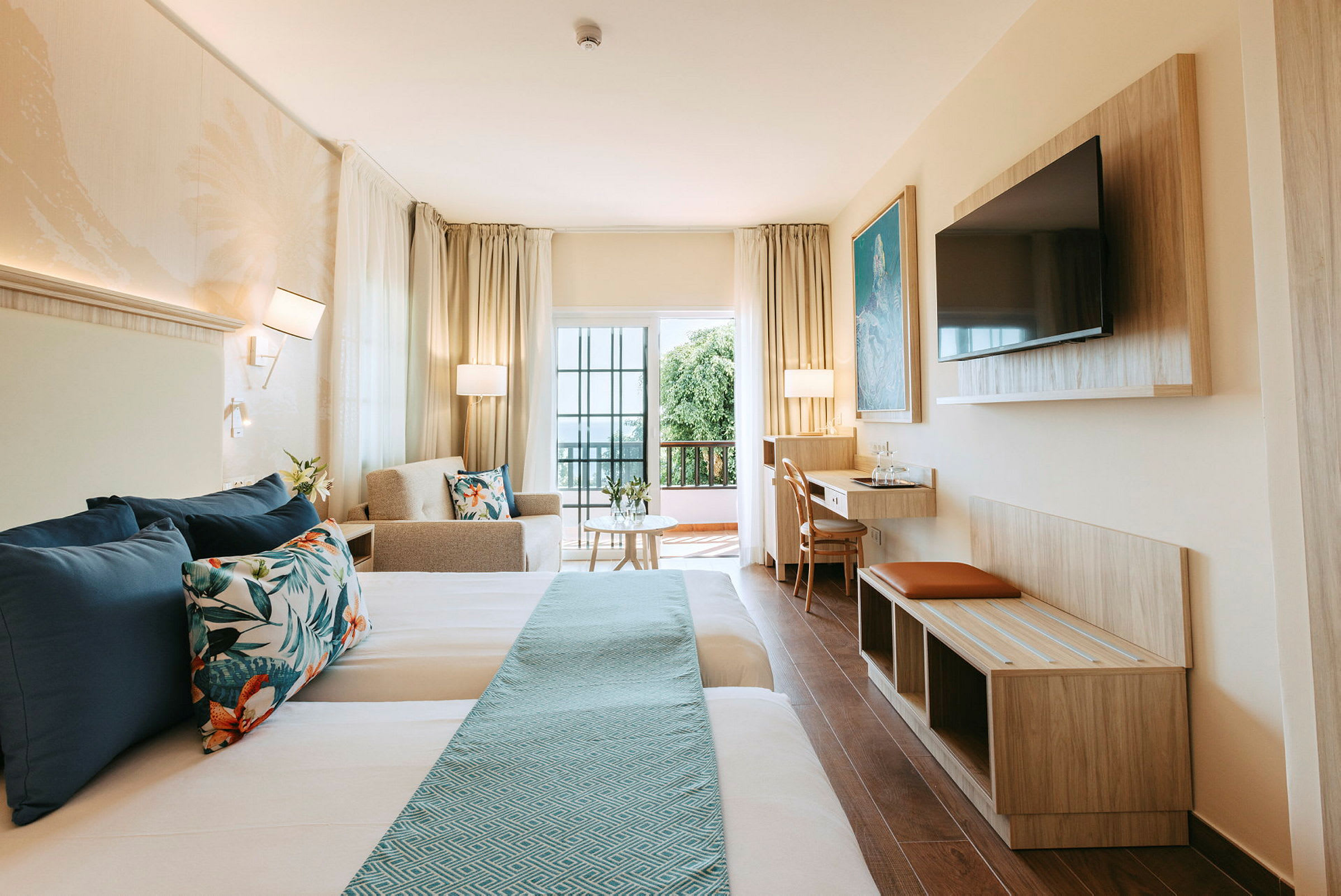 Et hotellrom med to senger, sofa, bord og TV, samt en balkong med glassdører. (AI-generert tekst)