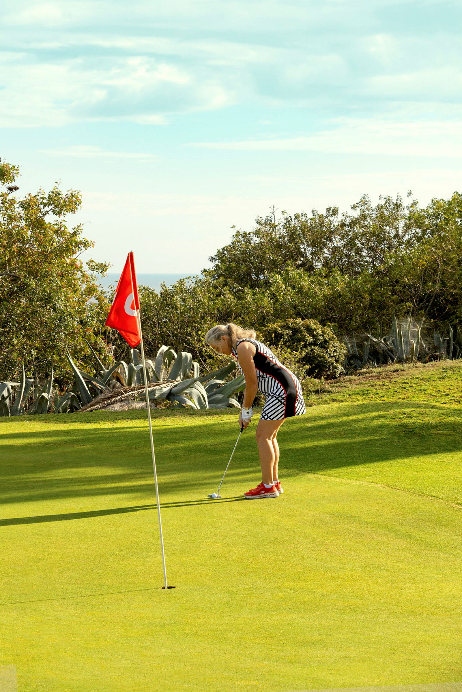 En person spiller golf og sikter mot hullet nær en rød flagg, med grønn natur i bakgrunnen. (AI-generert tekst)