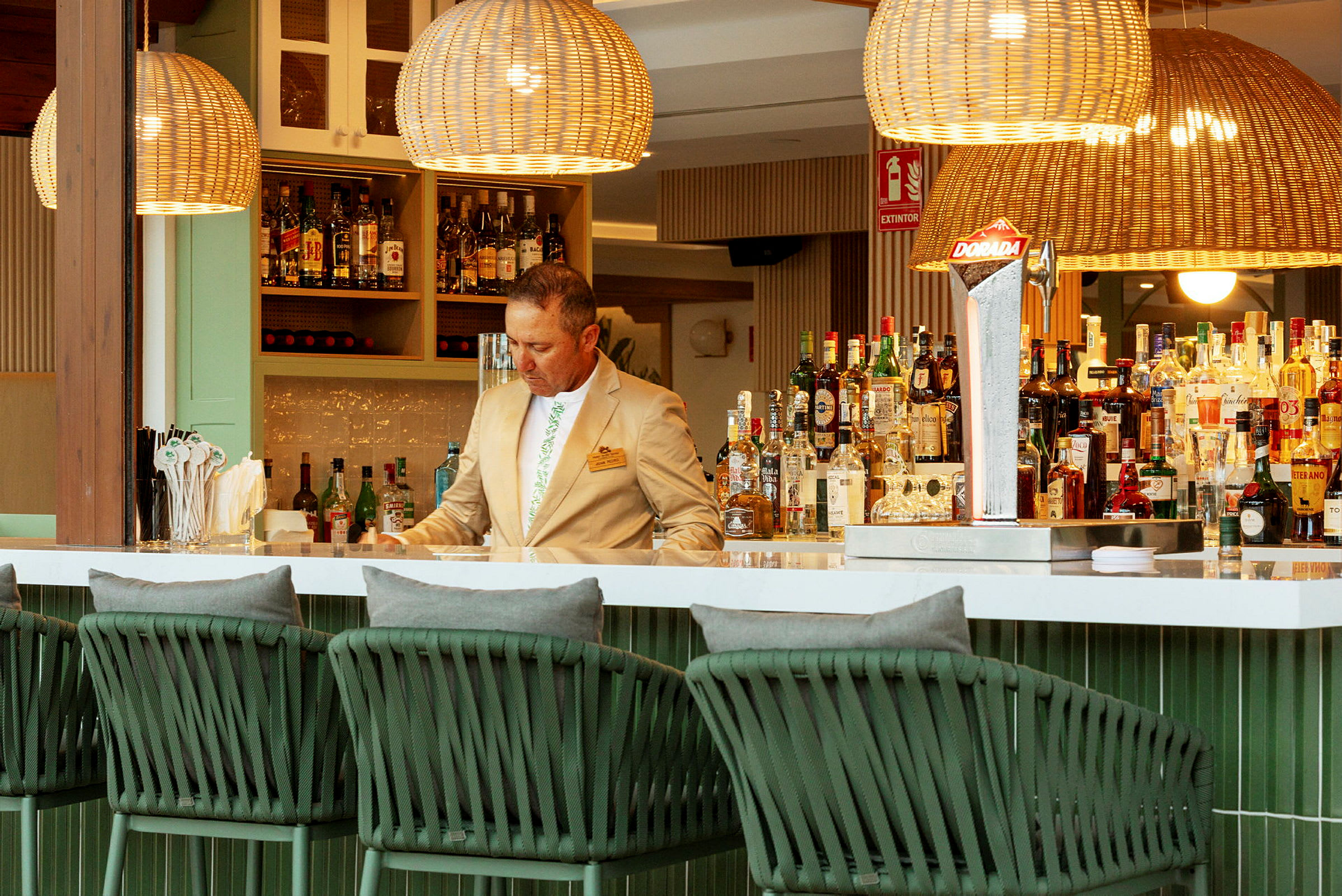 Interiør av en elegant bar med flettede lamper og stoler, samt et utvalg av flasker og drikker på hyllene. (AI-generert tekst)