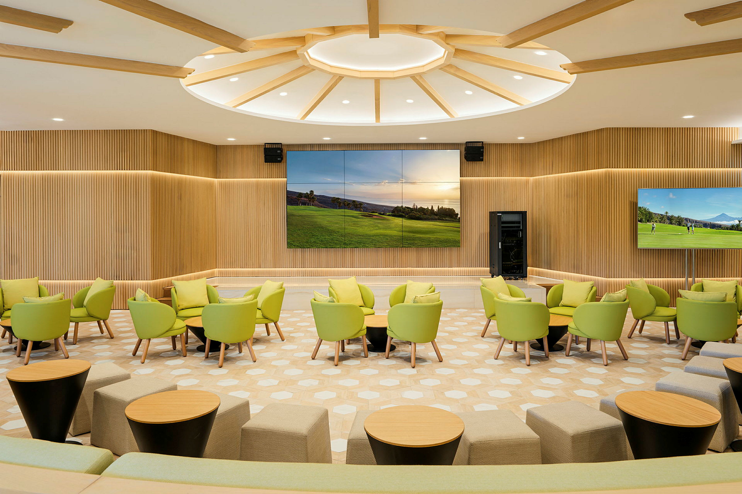 Et moderne møterom med grønne stoler, små runde bord og en stor skjerm på veggen omgitt av trepanel. (AI-generert tekst)