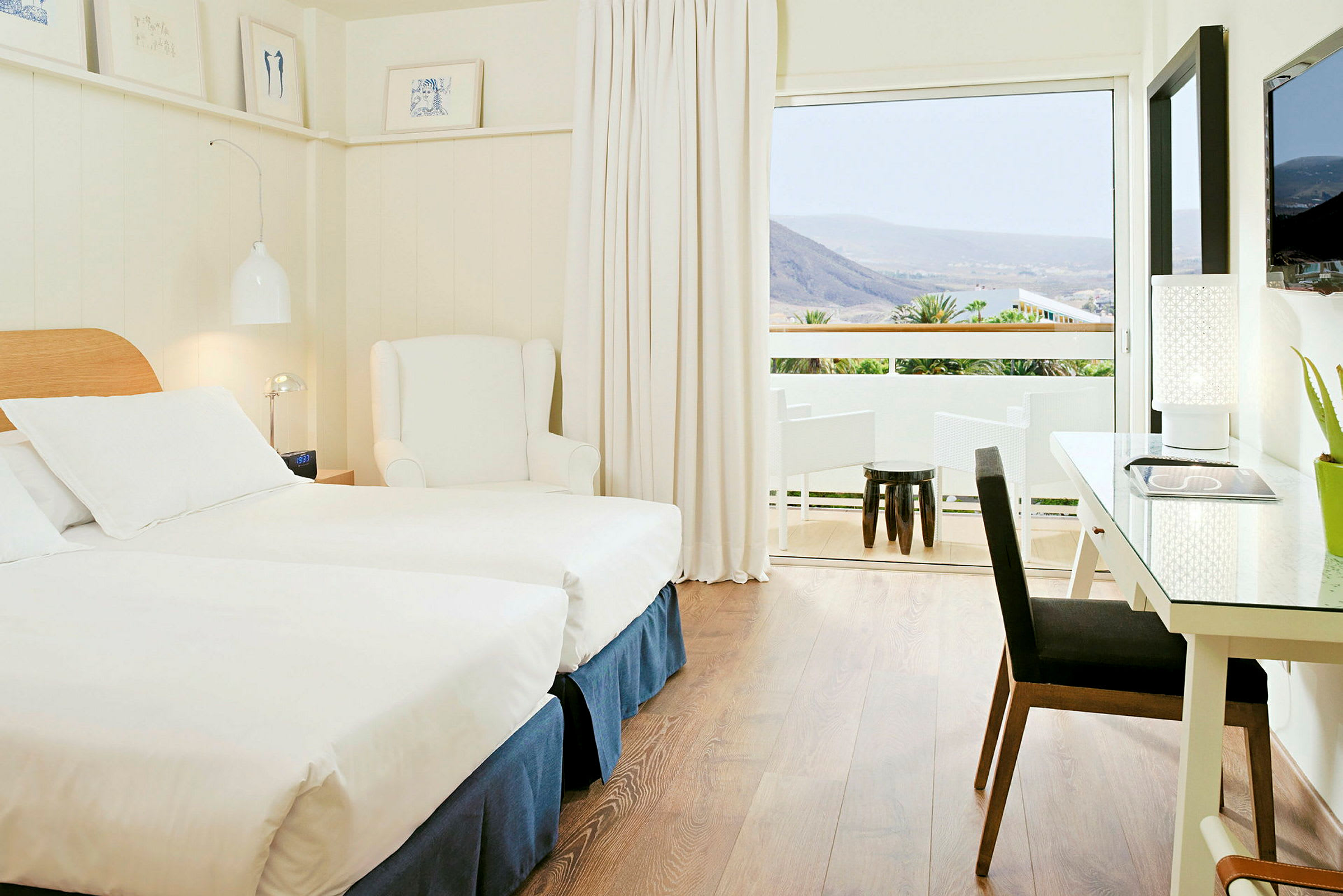 Et lyst og moderne hotellrom med to enkeltsenger, en hvit lenestol og et skrivebord med lampe. Et stort vindu fører ut til en balkong med utsikt over fjell og grønt. (AI-generert tekst)