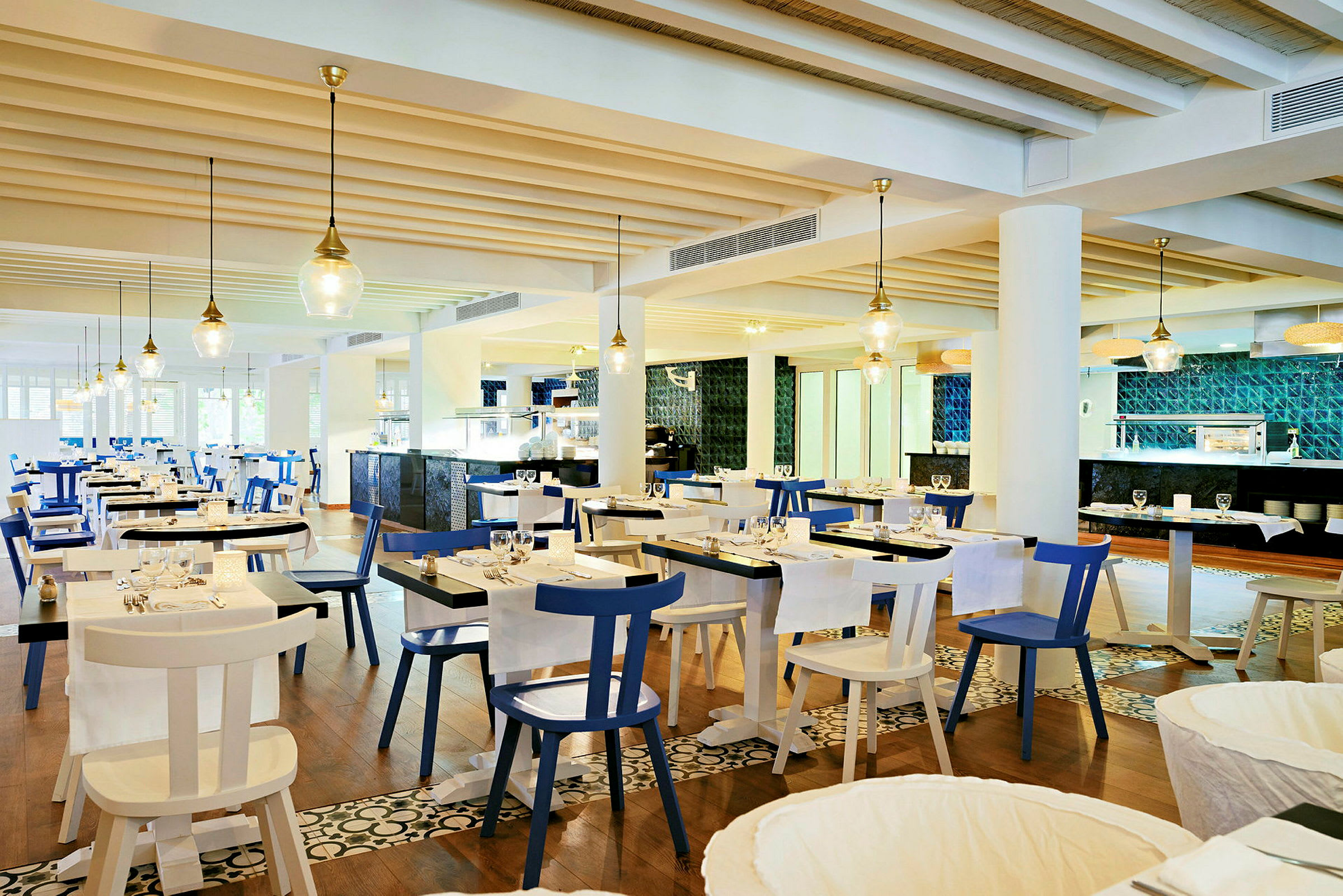 En lys og moderne restaurant med hvite og blå stoler og bord dekket for middag. Taket har synlige trebjelker og hengende lamper. Mønstret gulv og kjøkkenområde i bakgrunnen. (AI-generert tekst)