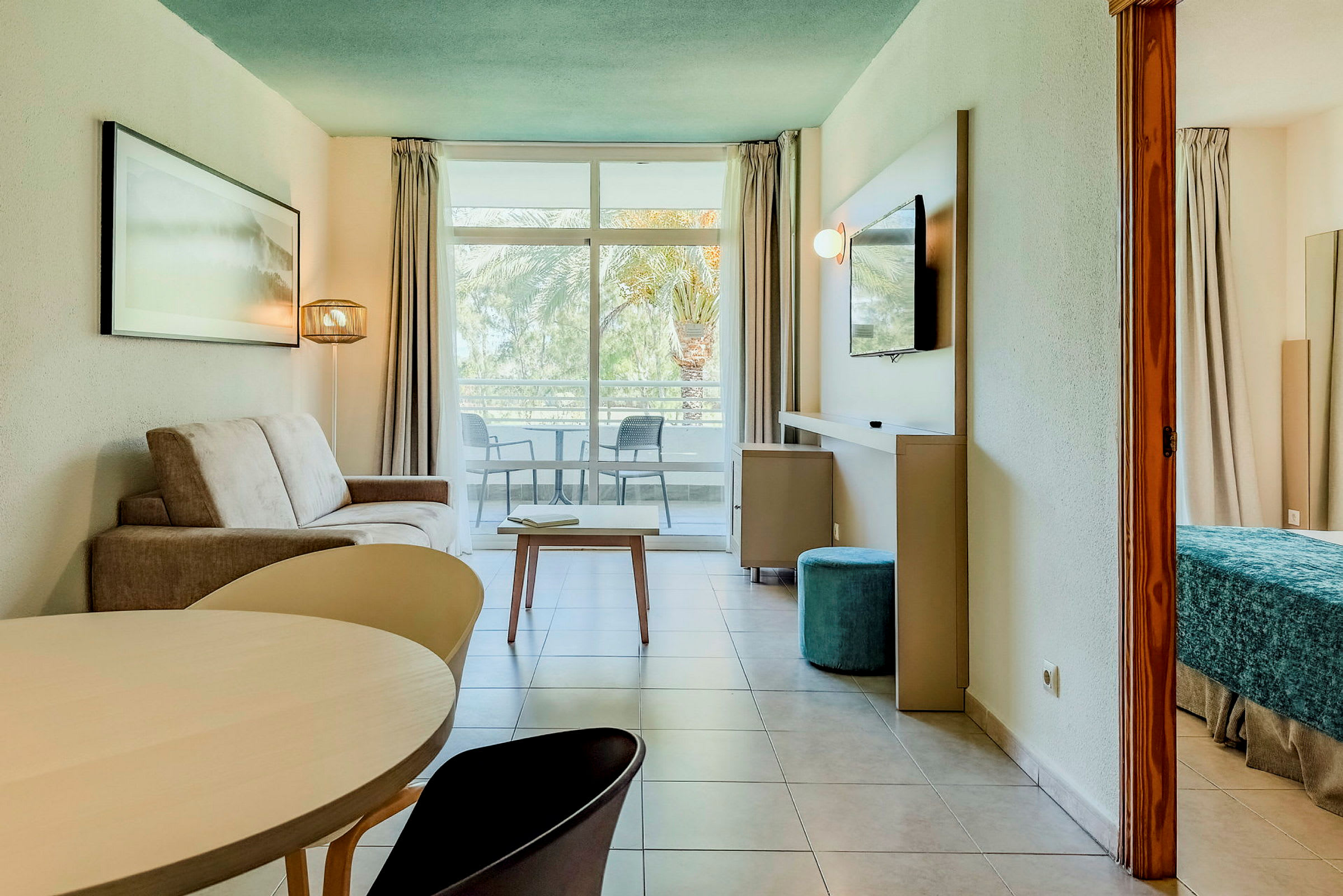 Moderne innredet hotellrom med sittegruppe, bord og soveområde. Balkongen har utsikt over palmer. (AI-generert tekst)