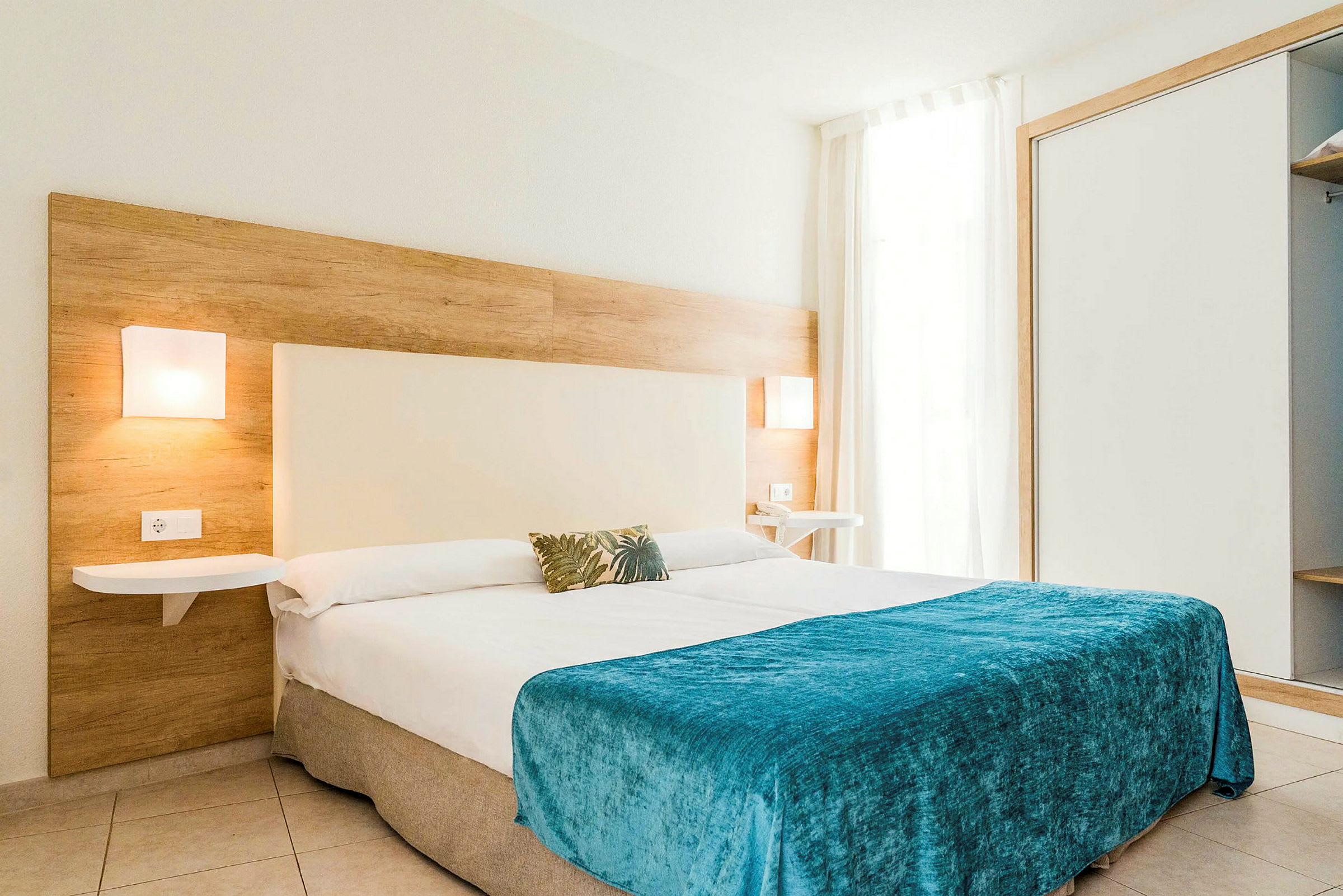 Moderne hotellrom med en stor seng dekket med et blått teppe og vegglamper på sidene. (AI-generert tekst)