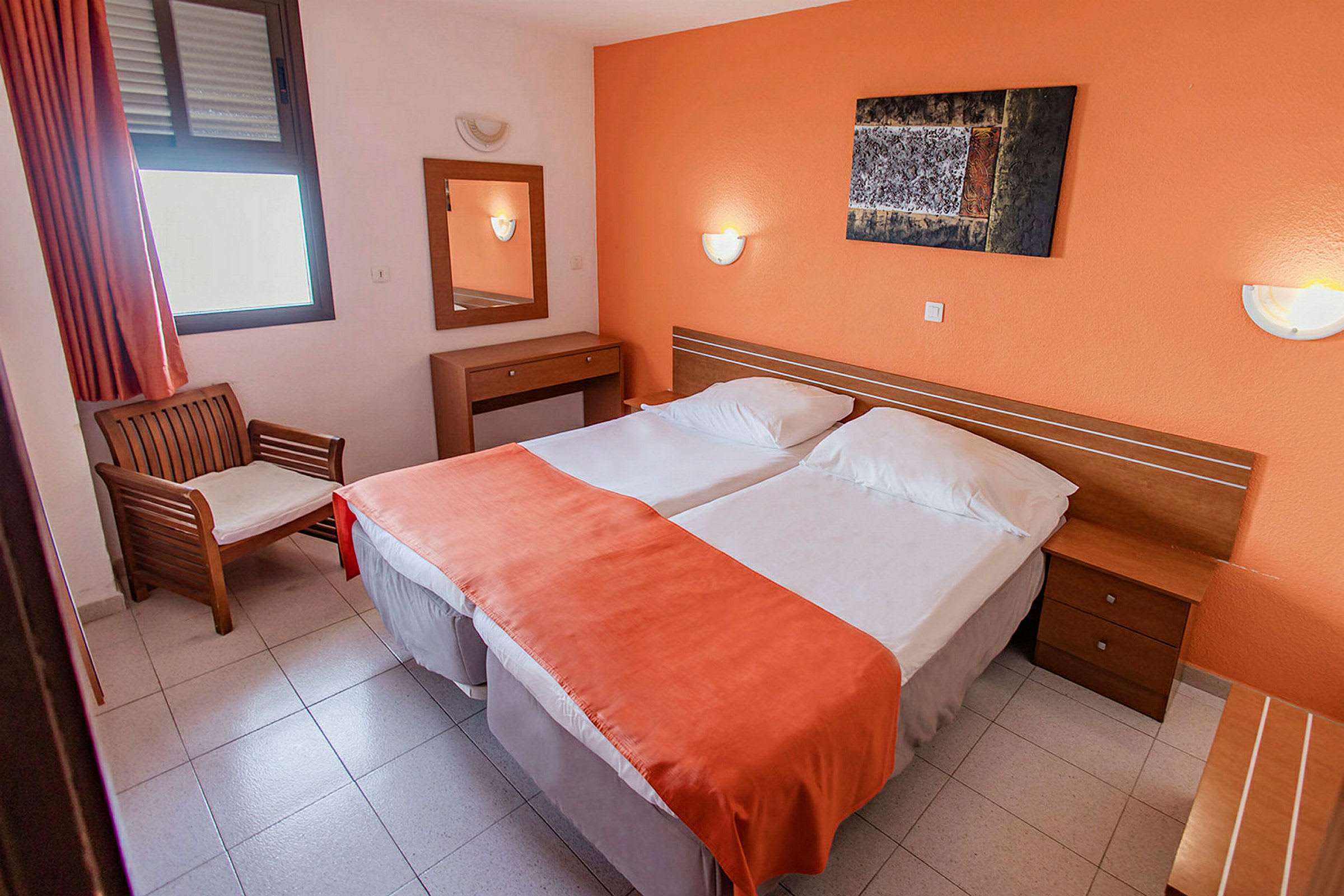 Et hotellrom med to enkeltsenger kledd med oransje sengeteppet, en stol ved siden av et vindu, og et speil med bord på veggen. (AI-generert tekst)