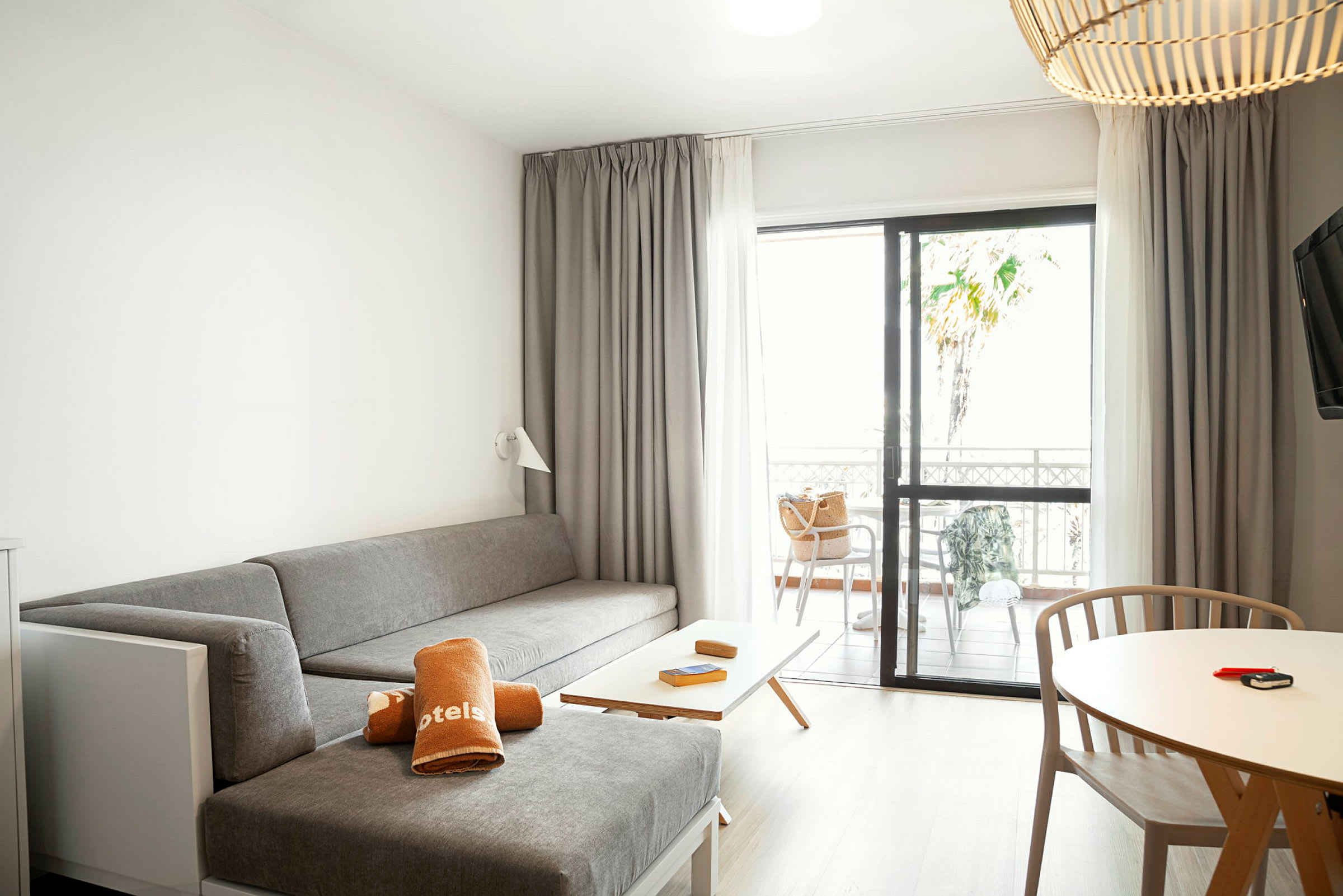 Et hotellrom med en grå sittegruppe, et bord med trestoler og en terrasse gjennom glassdører. (AI-generert tekst)