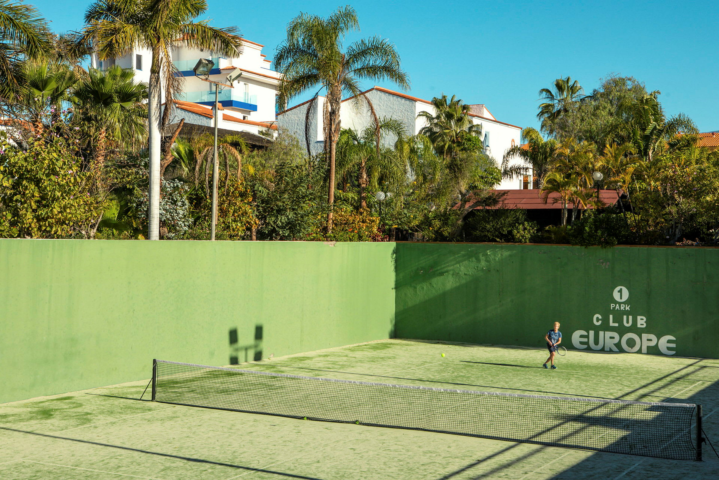En utendørs tennisbane med en person som spiller tennis, omgitt av grønne planter og bygninger. (AI-generert tekst)