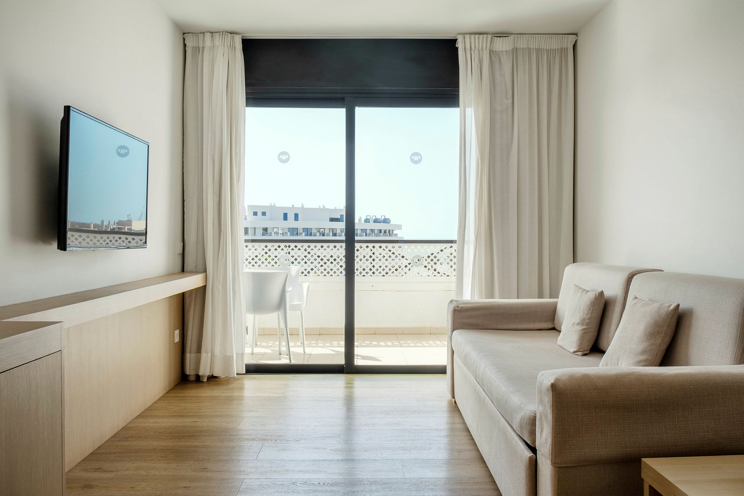 Et lyst og moderne hotellrom med sofa, veggmontert TV og stort vindu med utsikt mot en balkong. (AI-generert tekst)