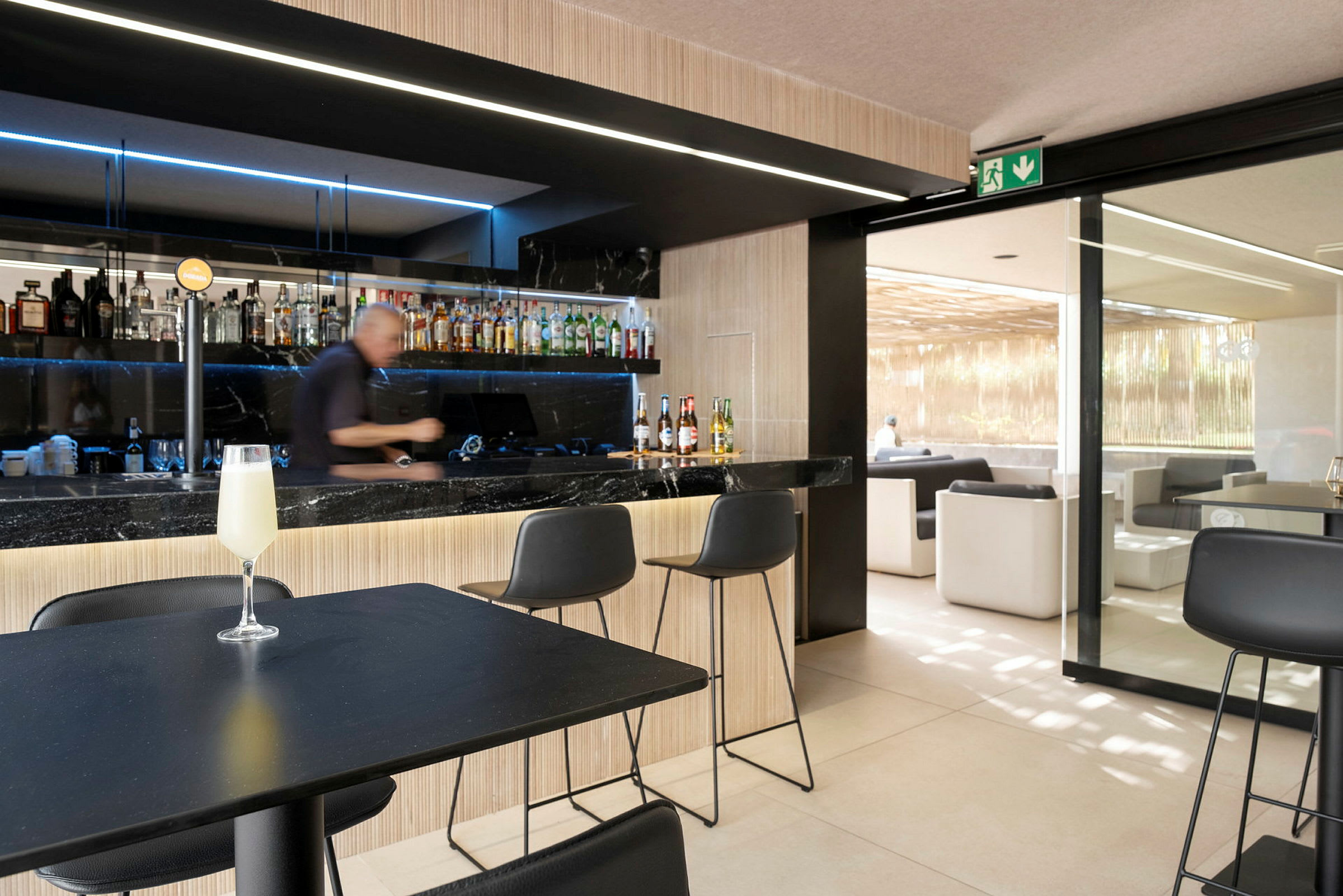 Et bilde av en bar med en bartender som forbereder drikkevarer. Foran ham står stoler, og en perlende drink er plassert på et bord. (AI-generert tekst)