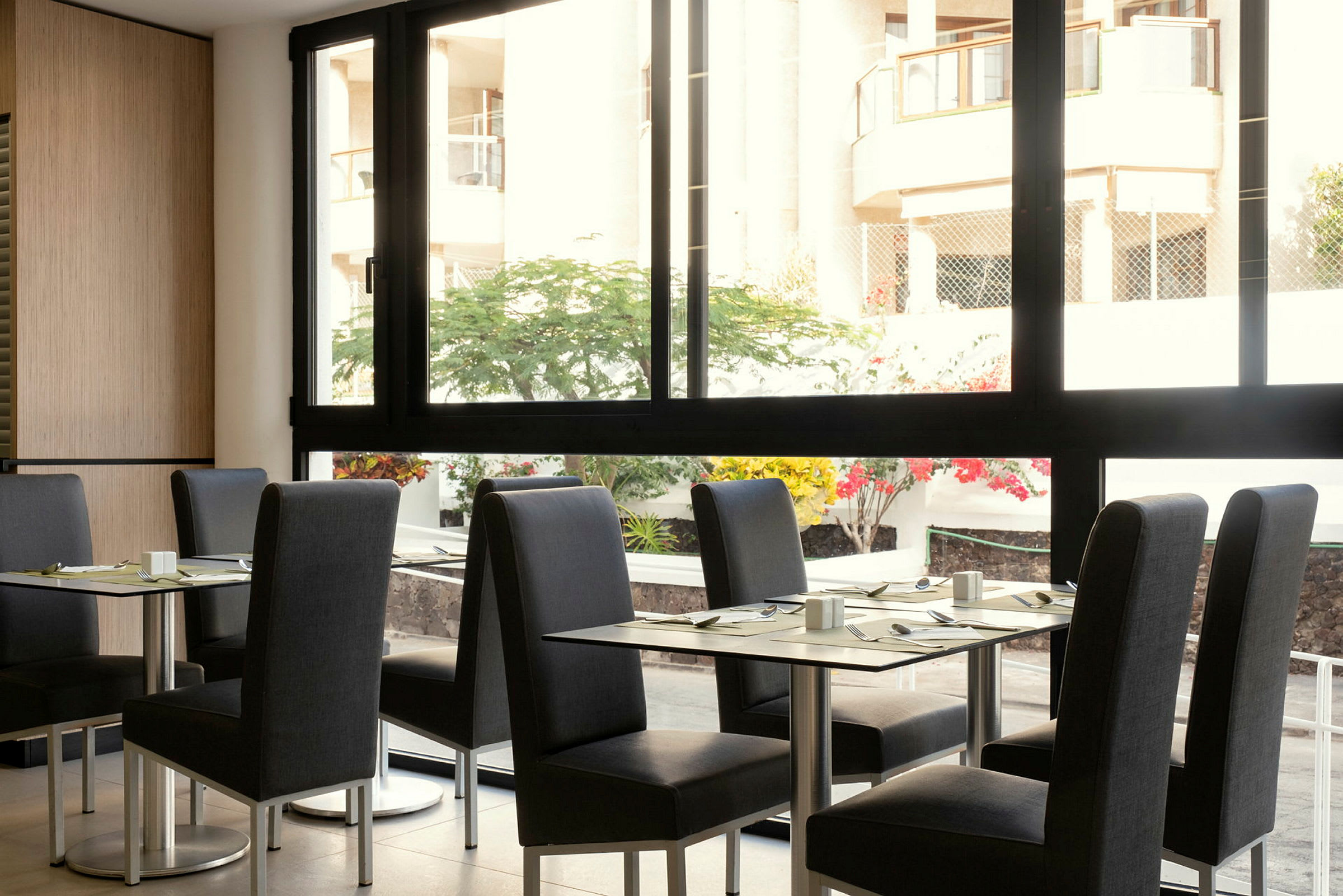En restaurant med bord og stoler, med utsikt til en gårdsplass gjennom et stort vindu. (AI-generert tekst)